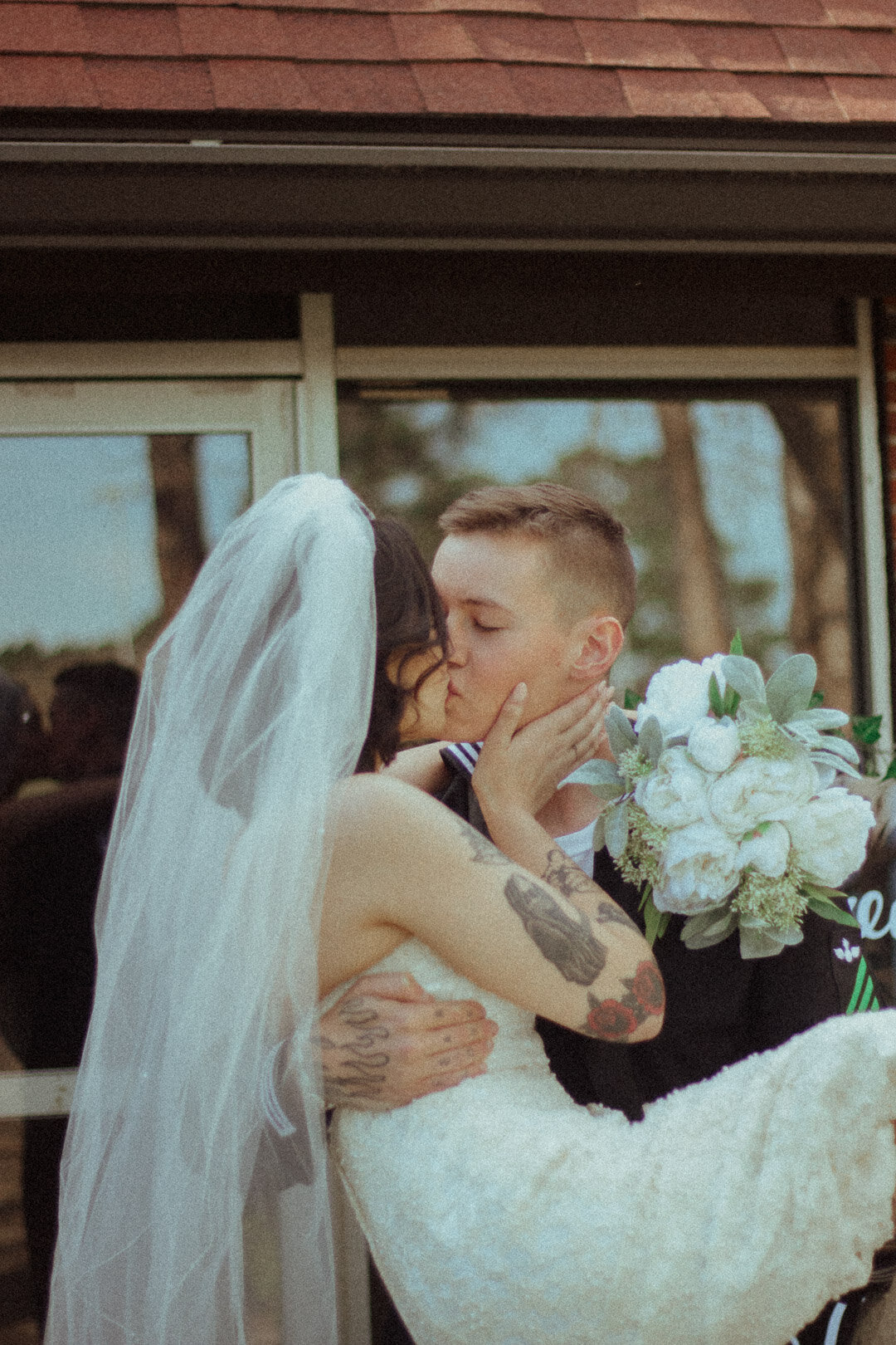 A couple, dressed in wedding attire, sharing a kiss during their wedding ceremony. The bride is wearing a white dress and veil, with tattoos on her arm. The groom is holding a bouquet of white flowers.