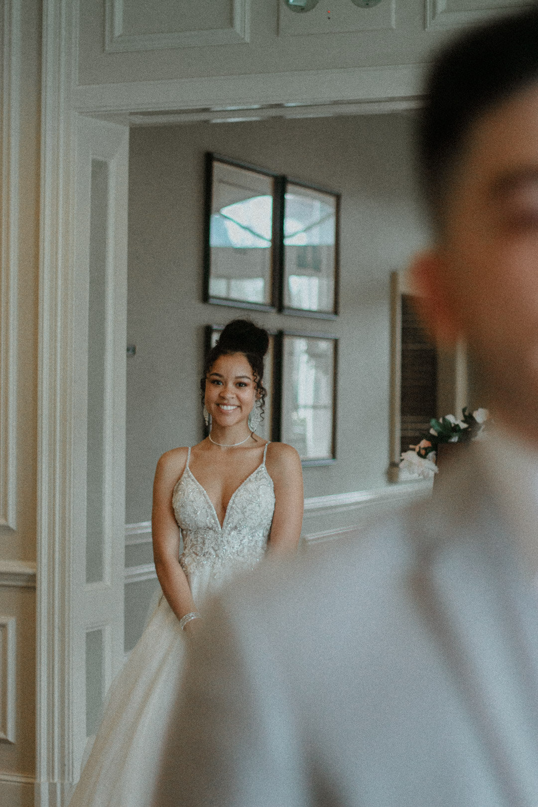 A bride in a white wedding gown smiles standing indoors near a doorway with windows.