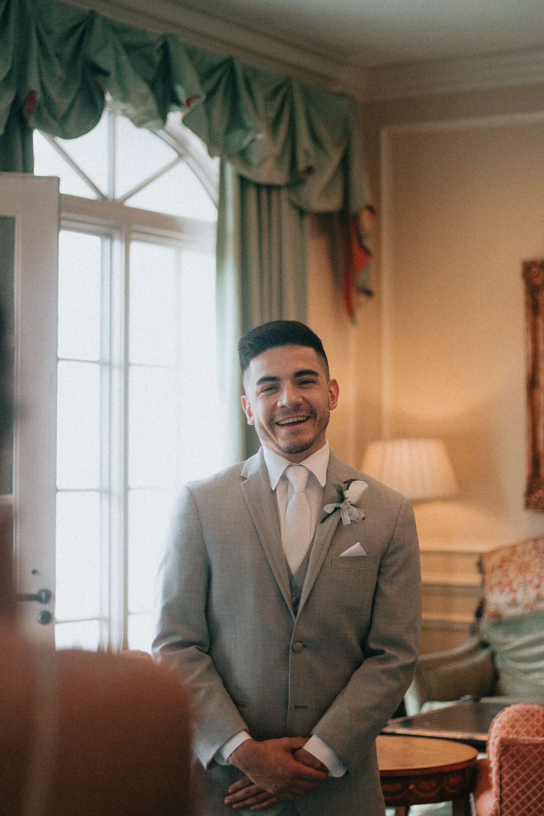 A smiling groom in a light gray suit and white tie, standing indoors near a window with green curtains, in a warmly lit room with classic decor.