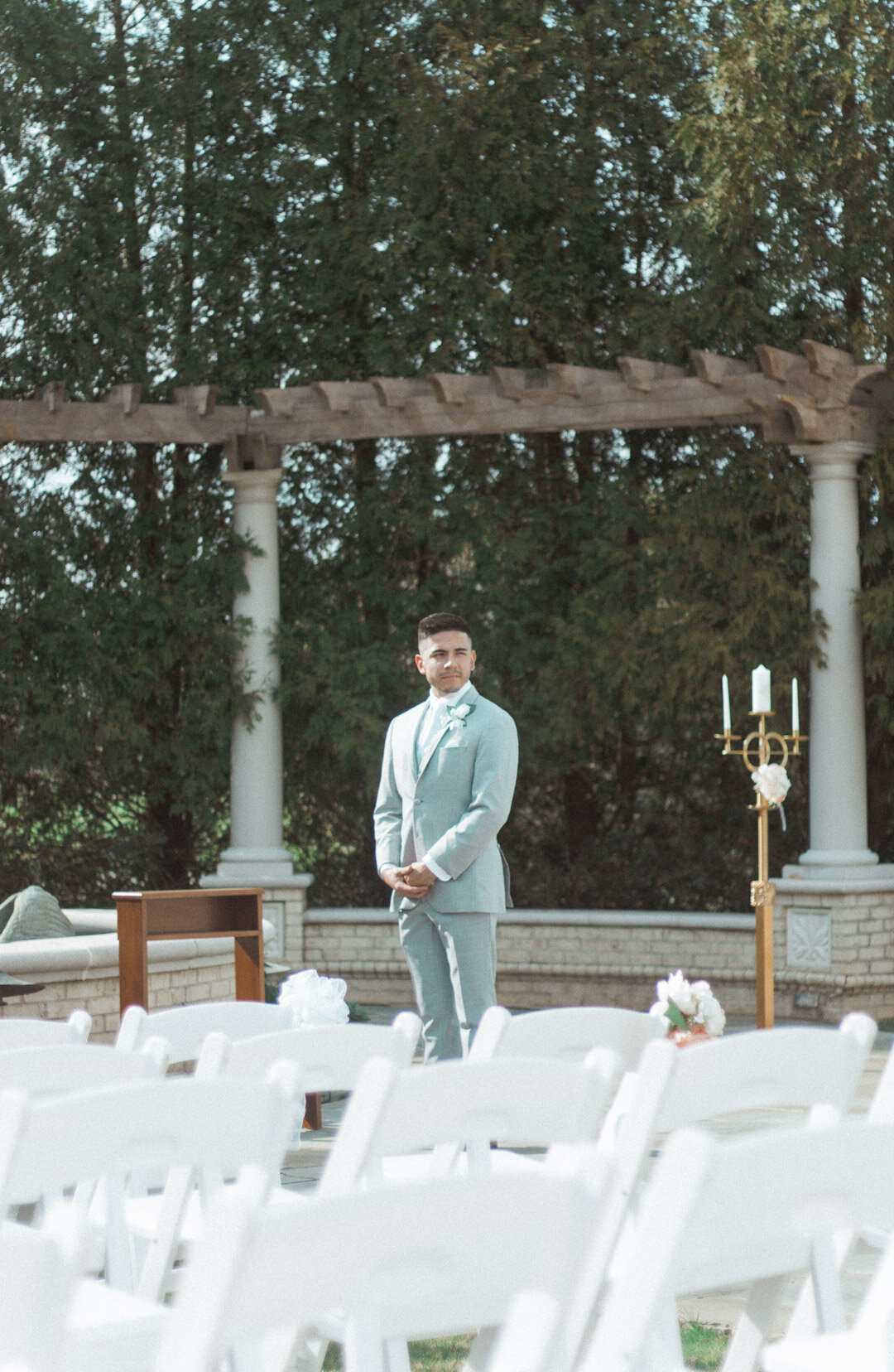 A groom in a light gray suit standing outdoors during a wedding ceremony, surrounded by white folding chairs and decorative candle holders, with greenery and trees in the background.