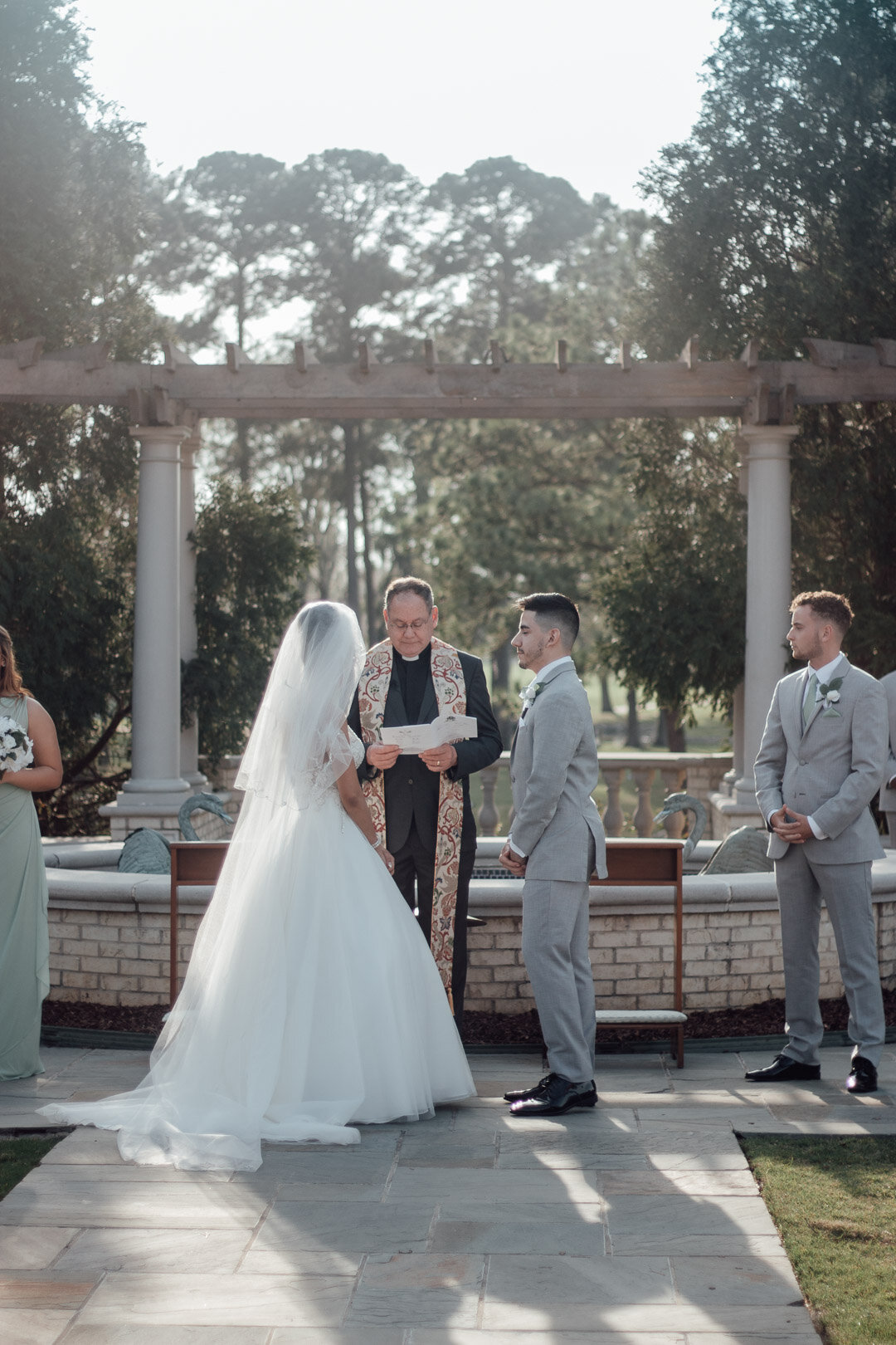A wedding ceremony outdoors with a couple standing before an officiant, surrounded by groomsmen and bridesmaids, in a garden setting with trees and a decorative stone fountain.