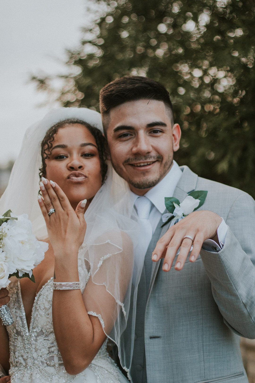 A newlywed couple outdoors, the woman with dark curly hair wearing a wedding dress, veil, and ring, showing her hand with the ring, and the man in a light gray suit with a boutonniere, smiling as they pose for a photo.
