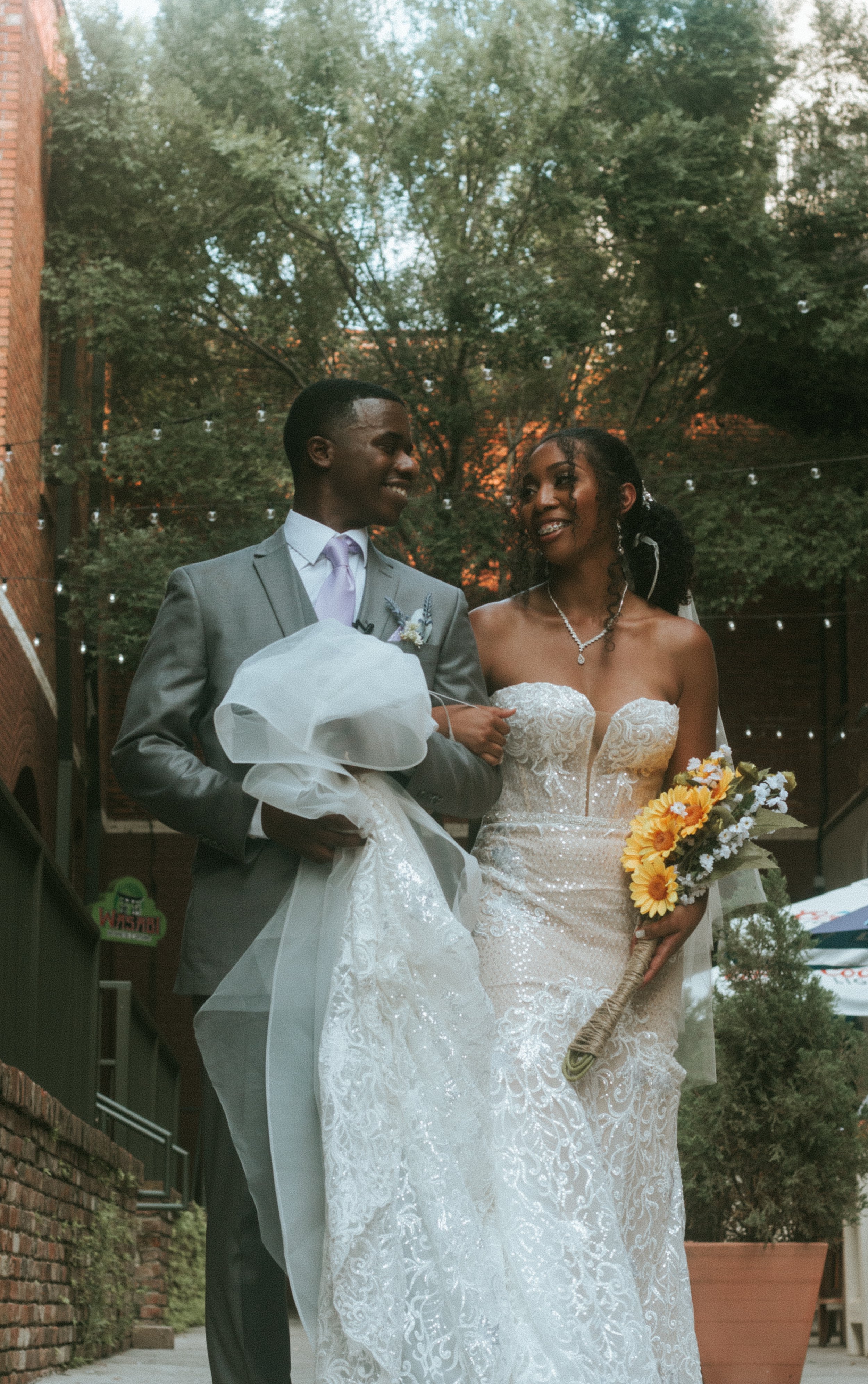 A bride and groom walking together at their wedding outdoors, smiling and holding hands, with trees and string lights in the background.