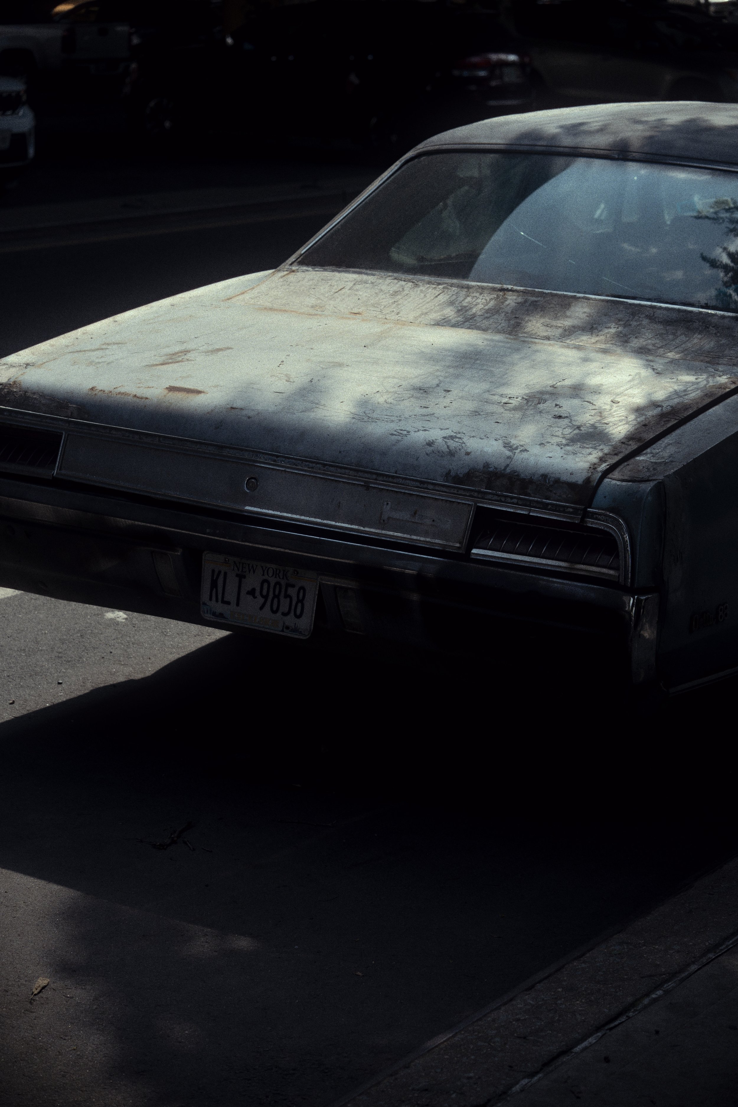 An old, dusty car with a weathered paint job parked on the street, with other cars visible in the background.