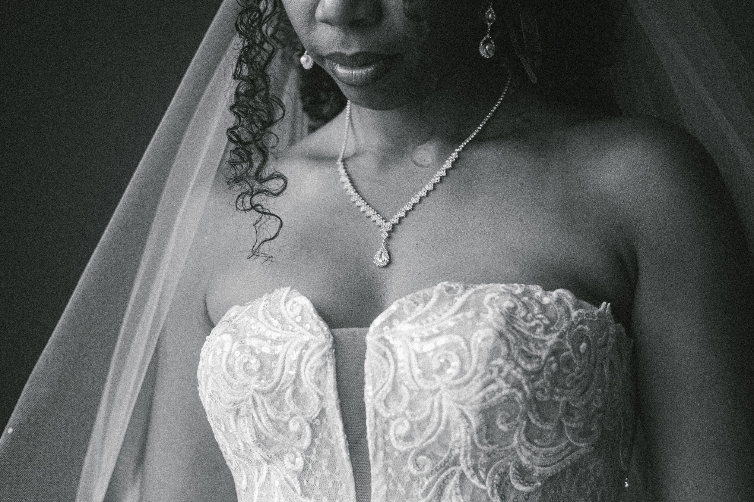 A close-up black and white photograph of a woman in a strapless, ornate wedding dress, wearing jewelry including earrings and a necklace, with her face partially visible.