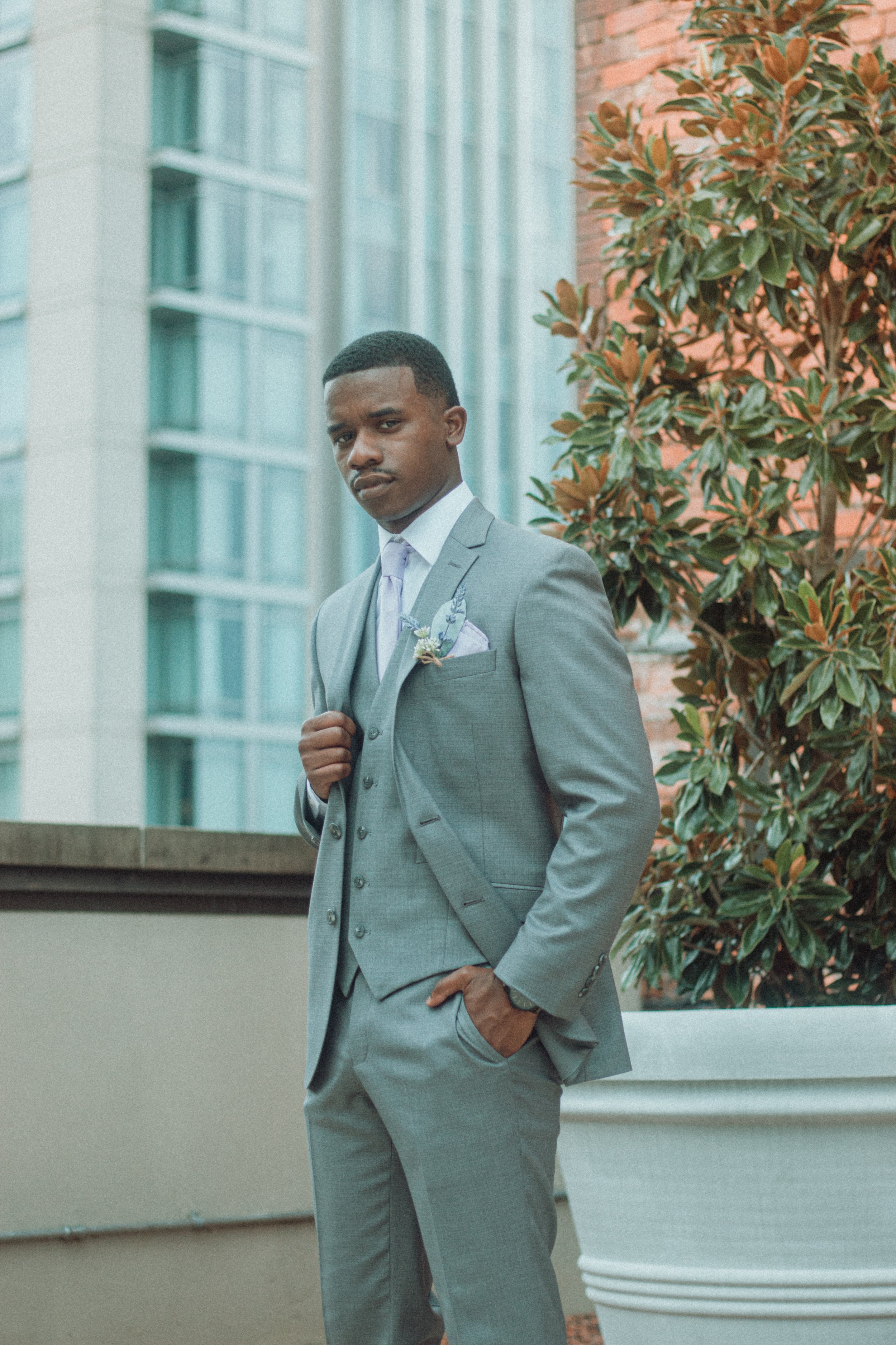 A man in a light gray suit with a purple tie and lavender pocket square posing outdoors near a building and large potted plant.