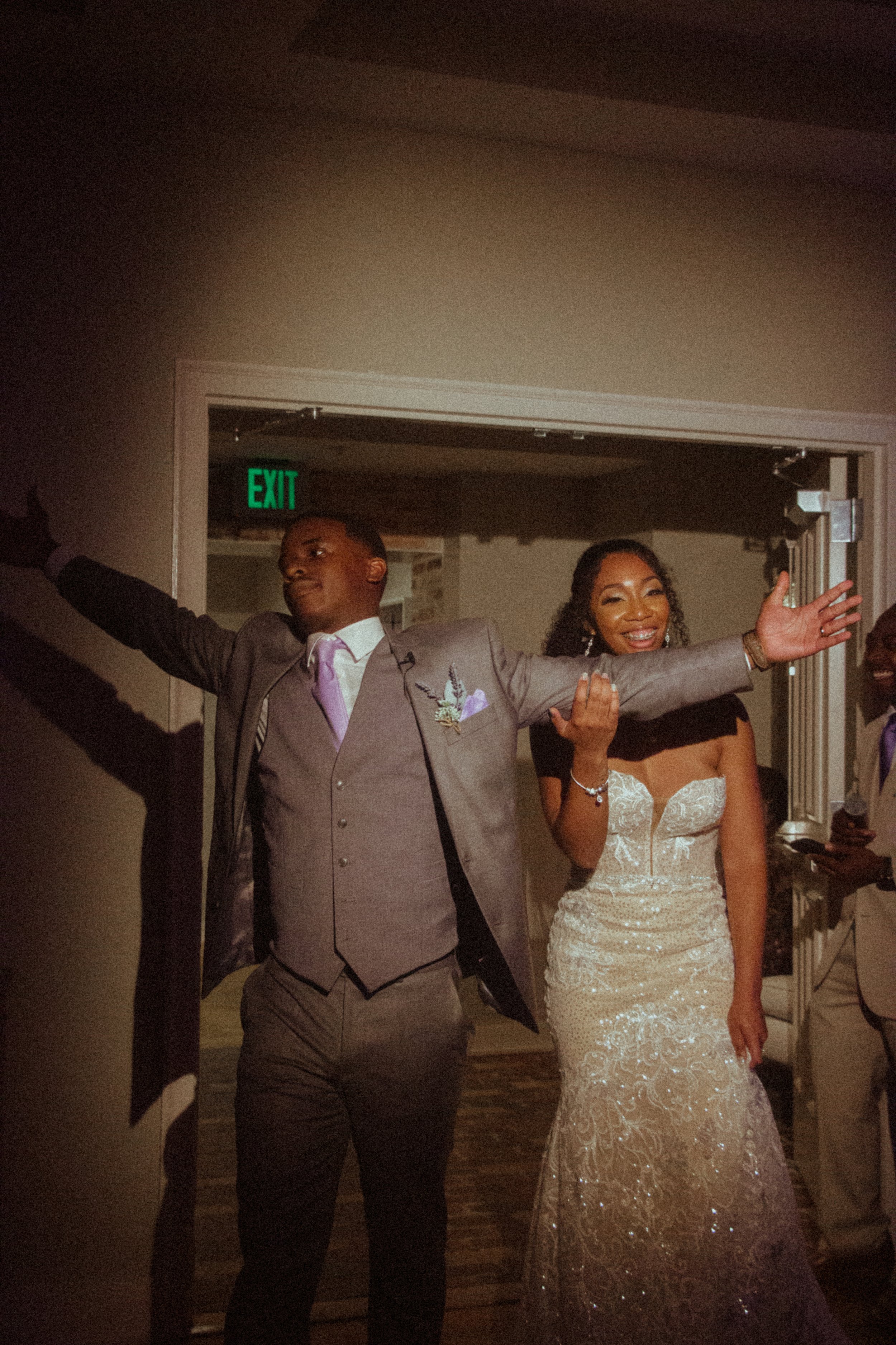 A joyful bride and groom celebrating at their wedding reception, with the groom with arms outstretched and smiling, and the bride smiling with her hand raised.