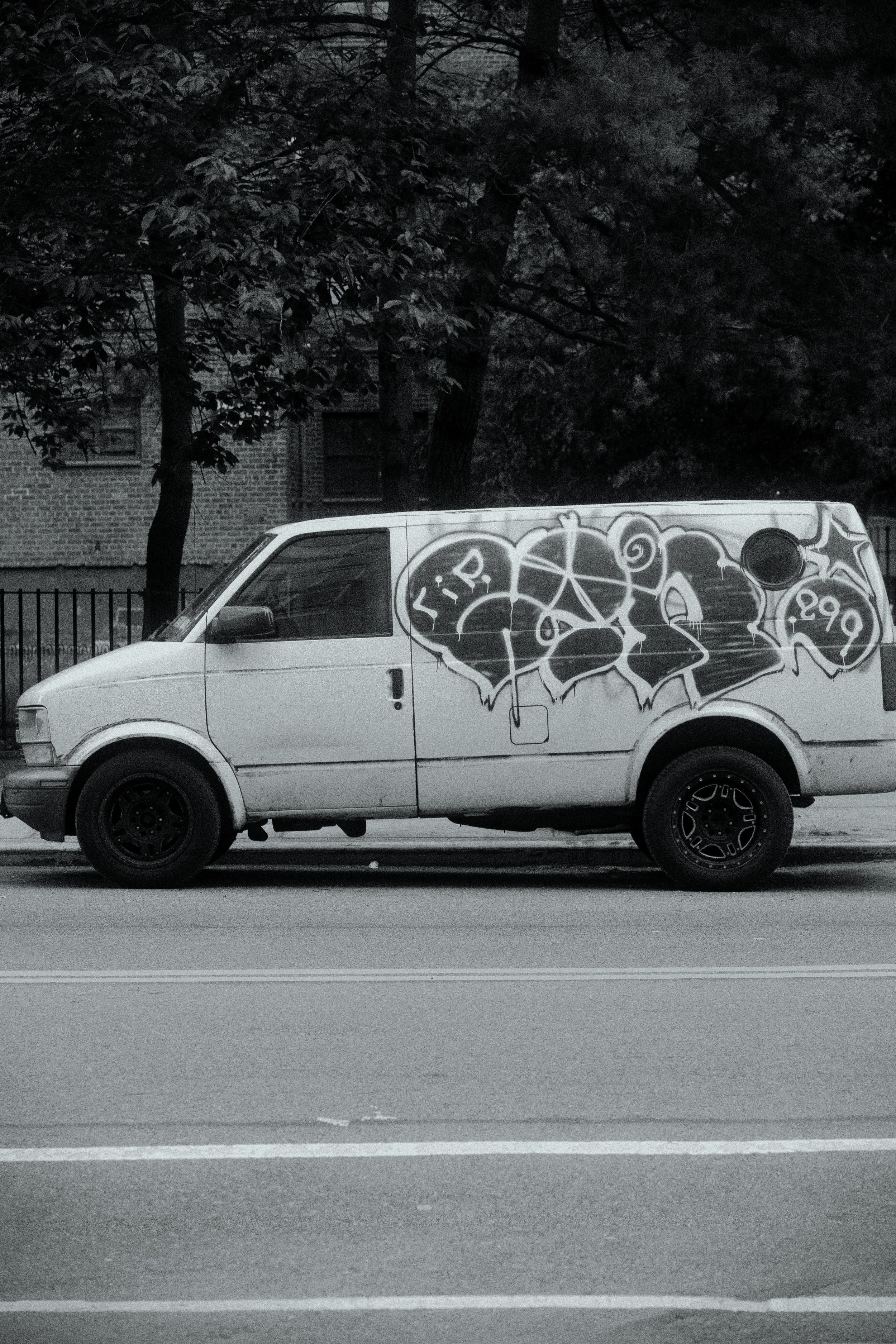 A white van parked on the side of a street with graffiti art on its side, featuring abstract shapes, symbols, and text, in a black and white setting.