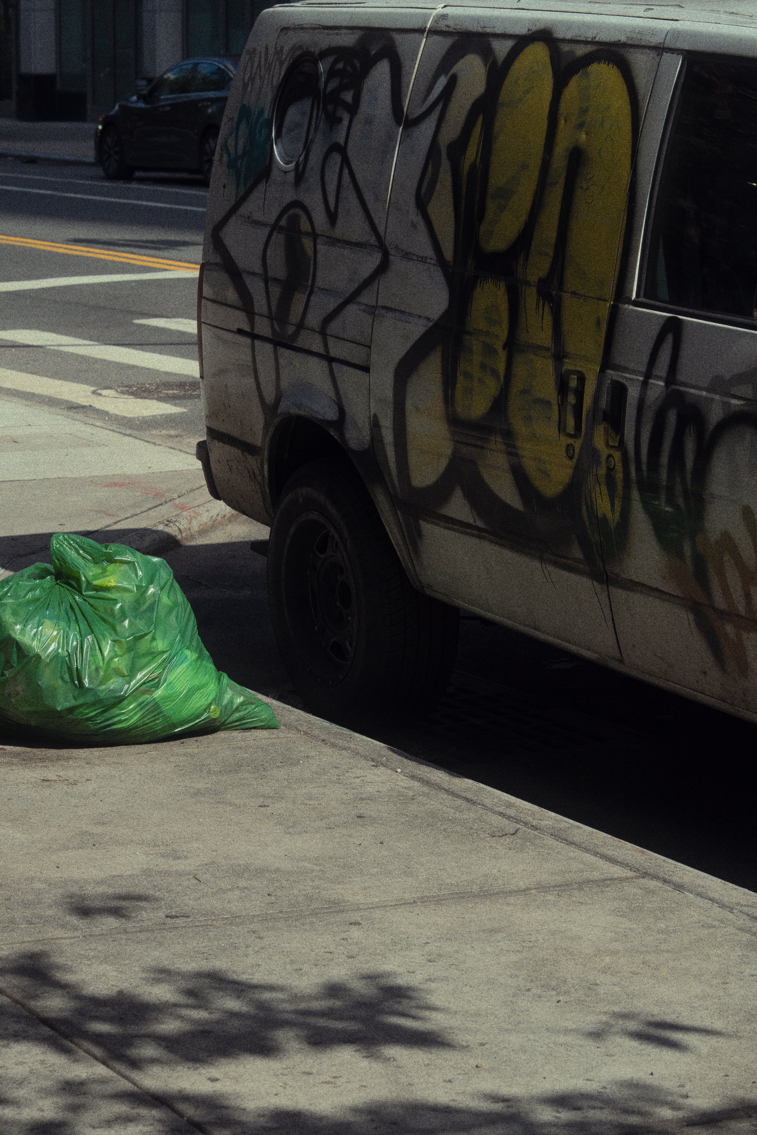 A green garbage bag on the sidewalk next to a graffiti-covered van parked on the street.