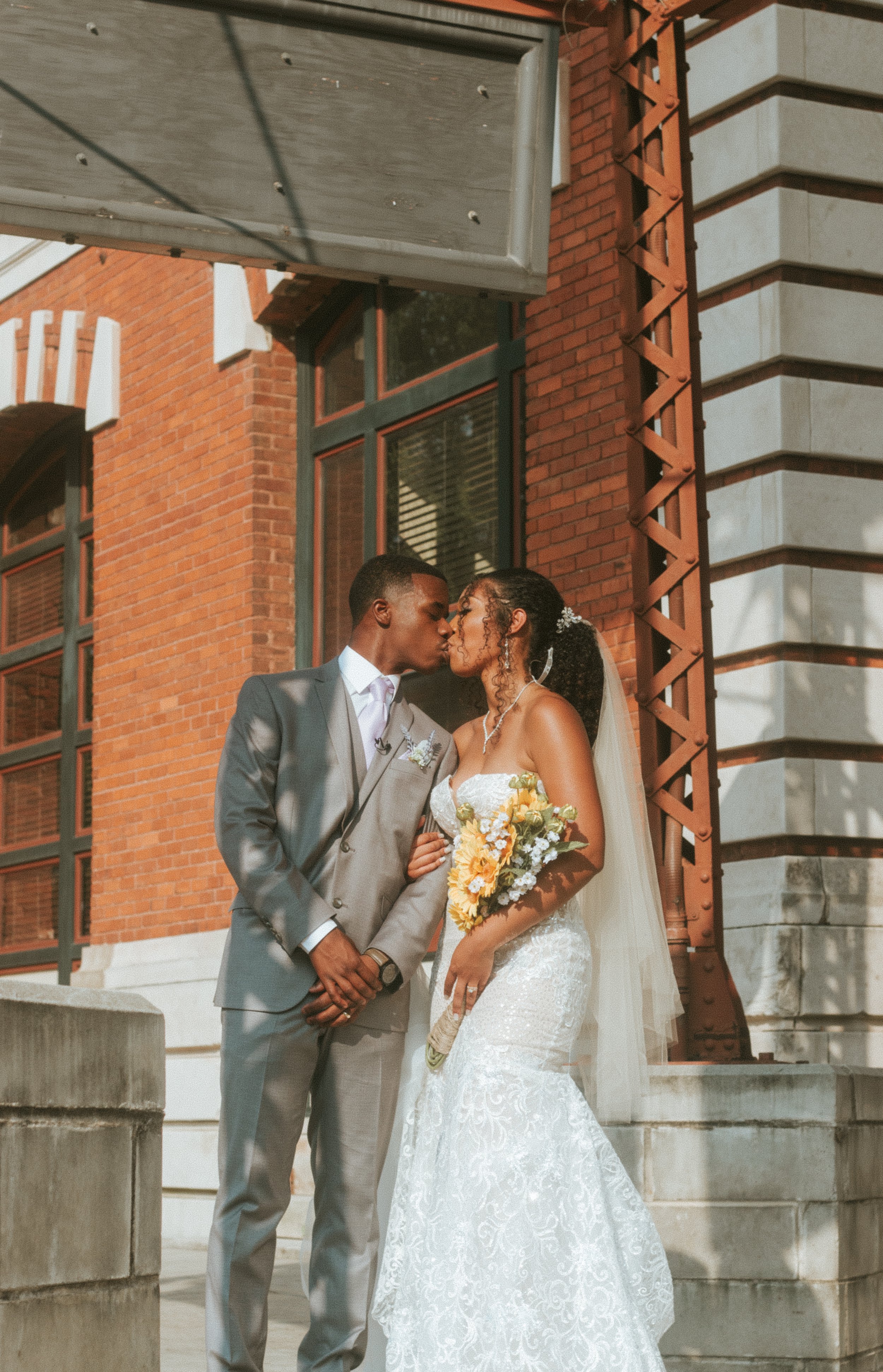 A bride and groom kissing outside a brick building on their wedding day. The bride is holding a bouquet of yellow and white flowers, and they are standing near concrete steps.