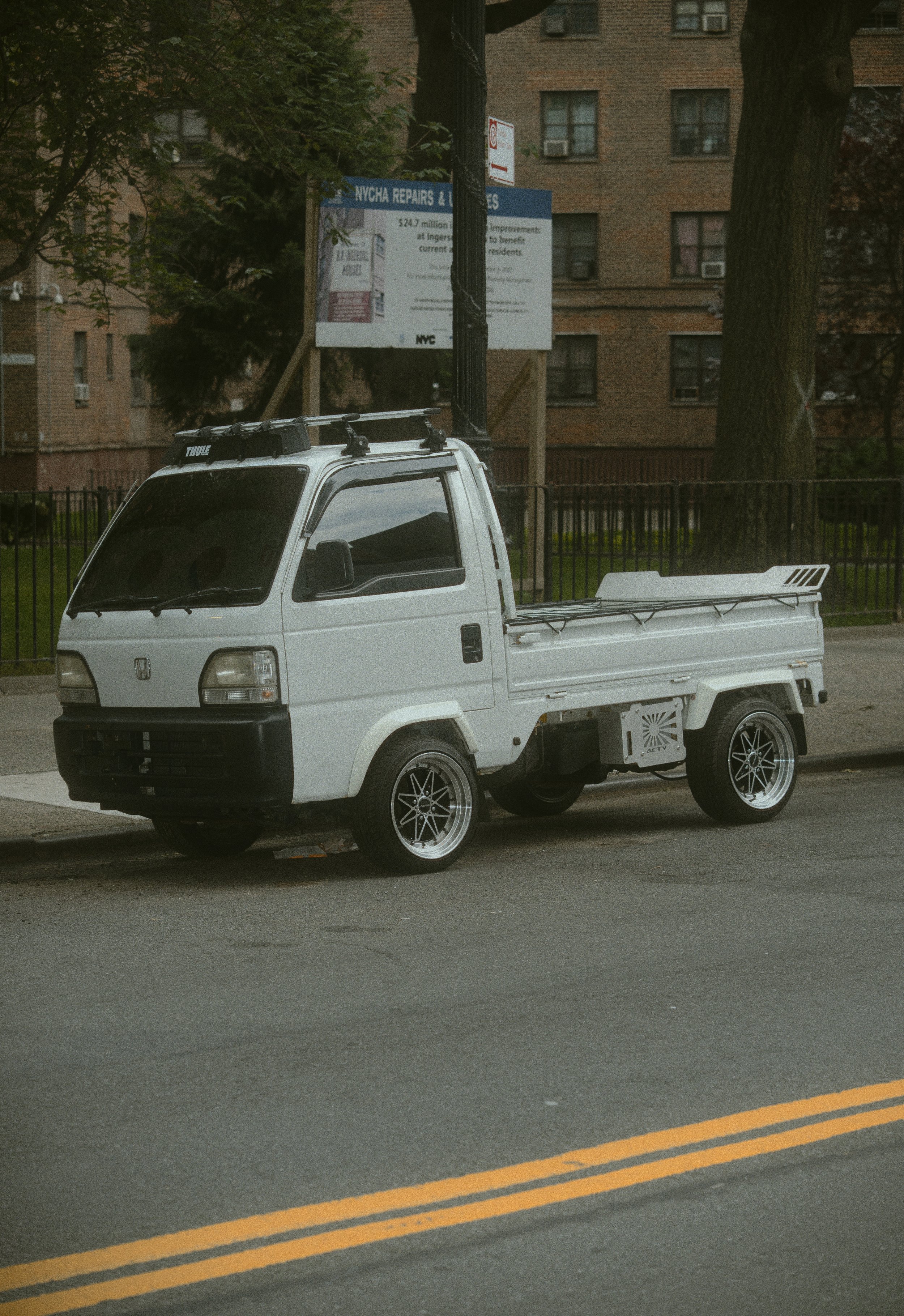 A small white pickup truck parked on the street near a sidewalk, with a black fence and brick apartment buildings in the background.