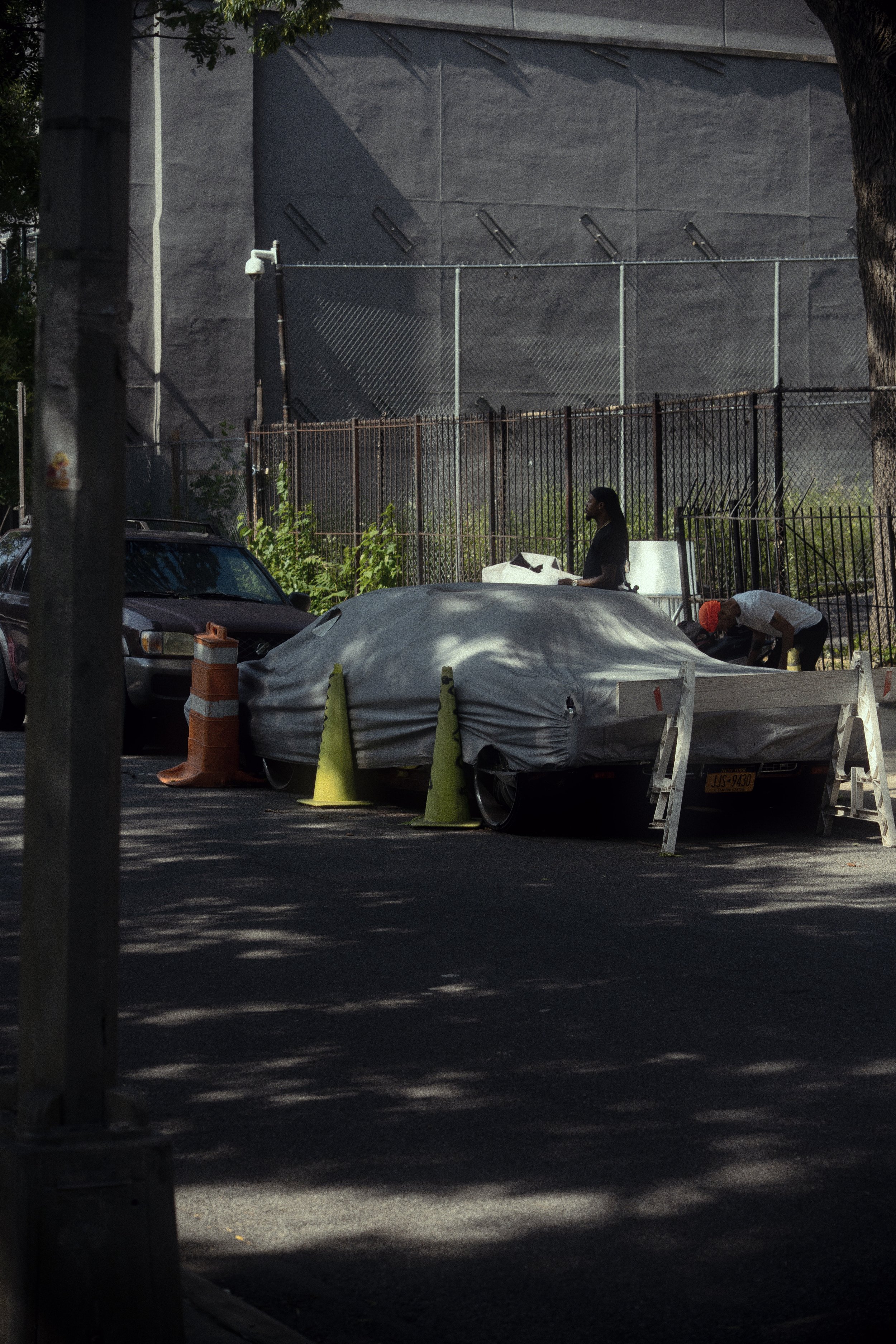 A car covered with a gray tarp parked on a city street, surrounded by traffic cones and barriers, with two people working nearby and a chain-link fence in the background.