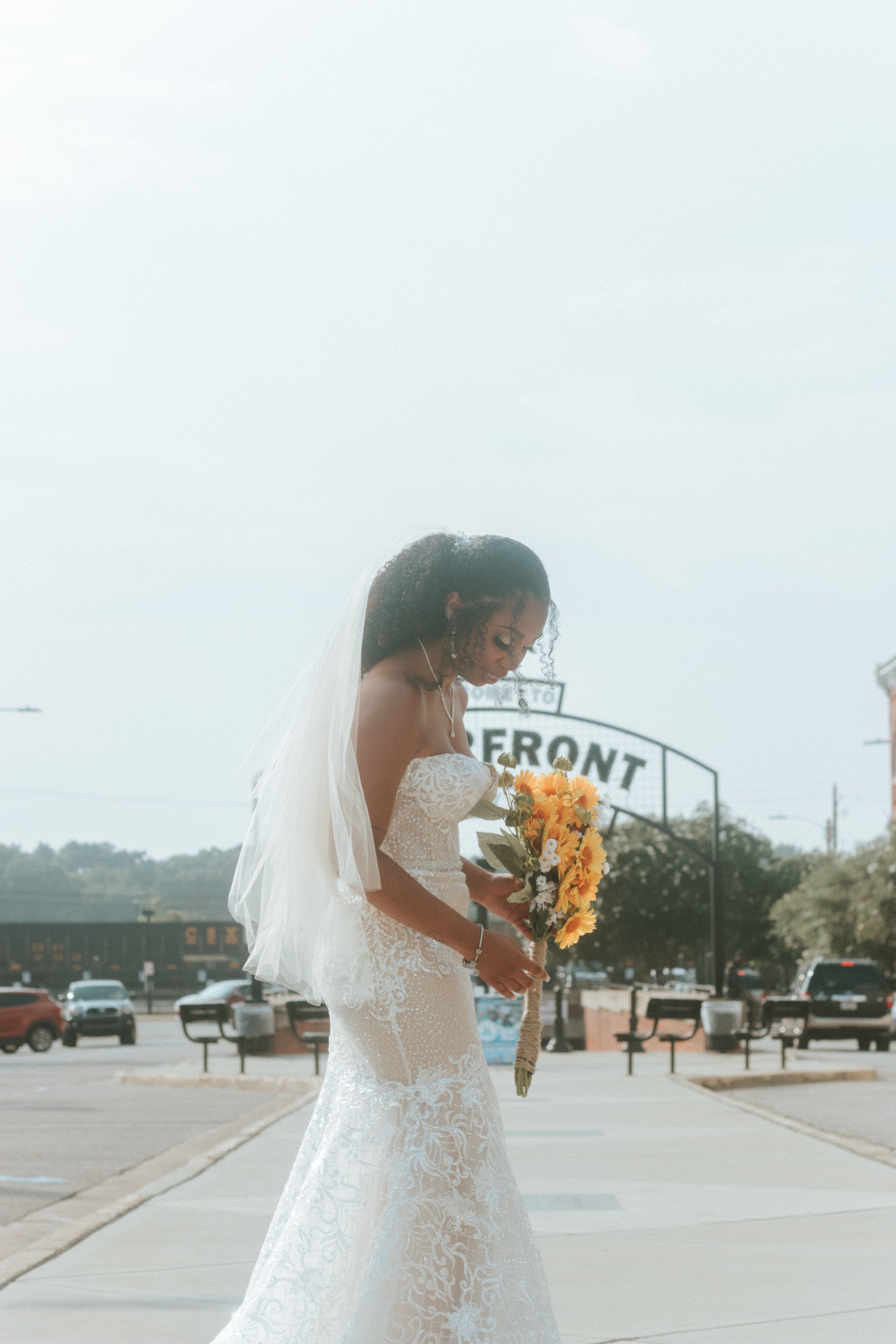 A bride in a white wedding dress holding a bouquet of yellow sunflowers, walking outdoors near a sign that says 'FERRONT' on a cloudy day.