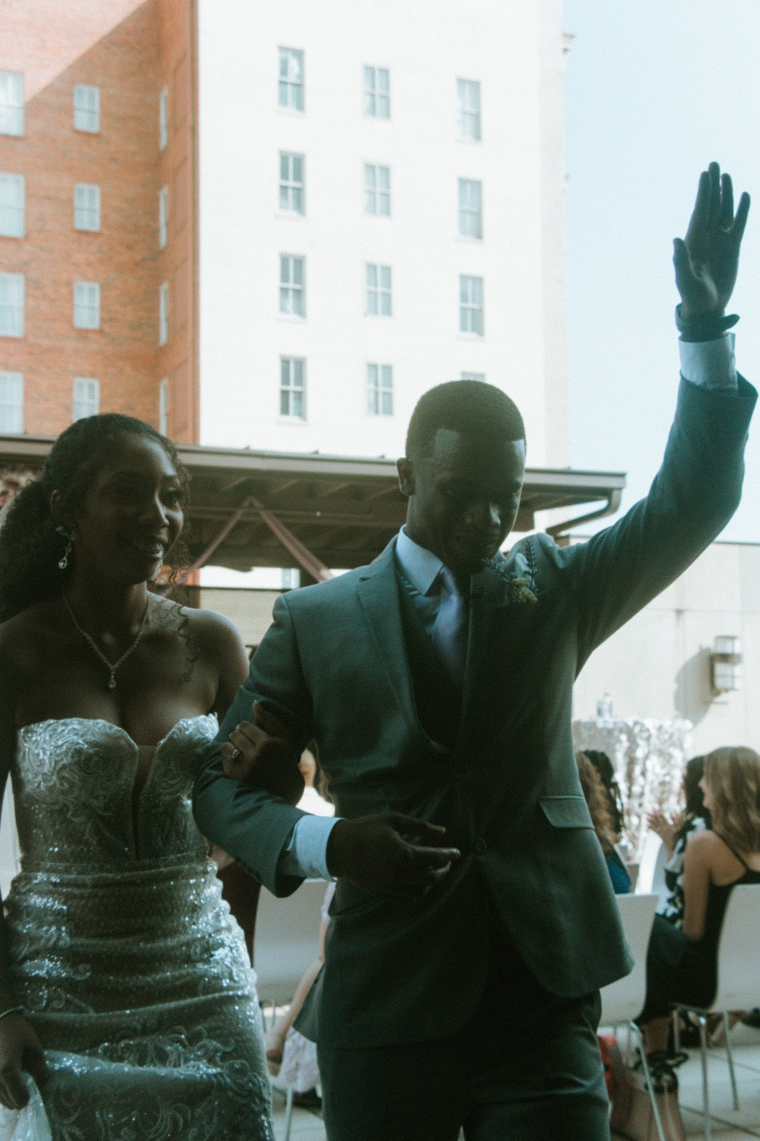 A couple at a wedding reception, with the man waving and the woman smiling, both dressed in formal attire.
