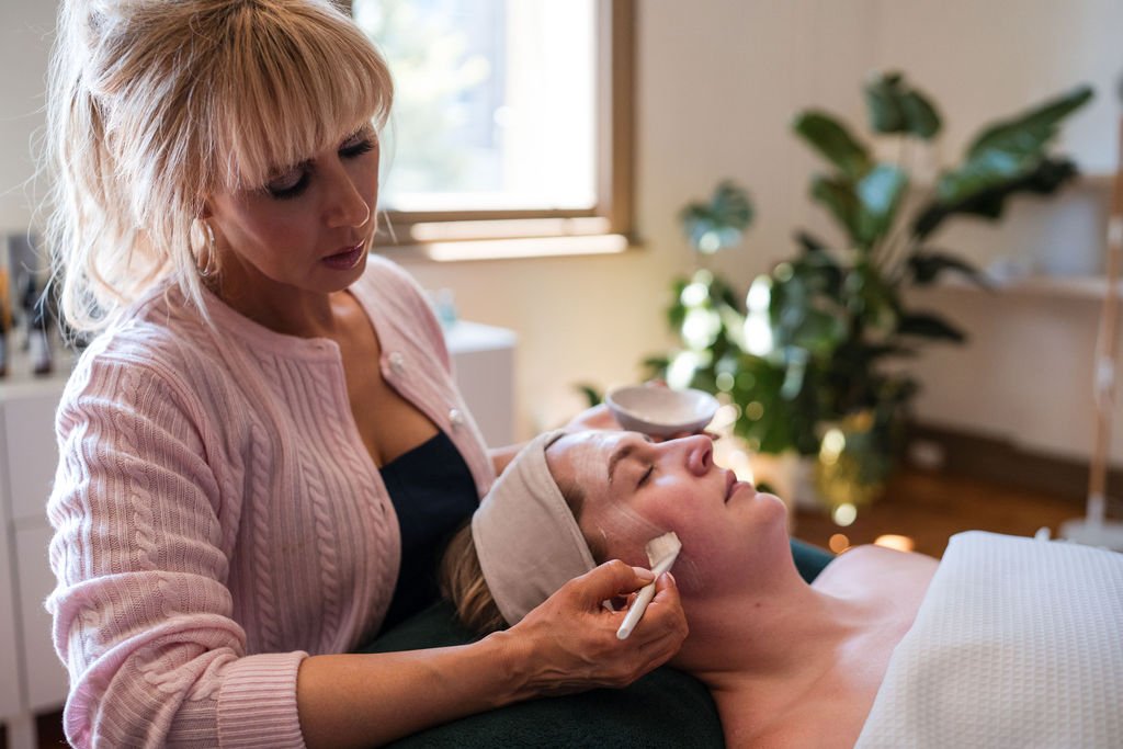 A woman is giving a facial treatment to a woman lying down with a towel over her chest, in a spa or beauty clinic, with a large plant in the background.