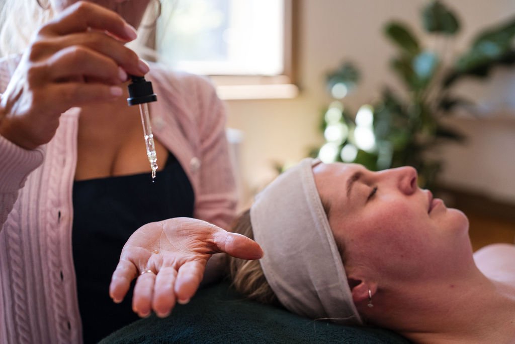 A woman lying down with her eyes closed, receiving an acupuncture treatment from a practitioner holding a dropper over her arm in a wellness setting.