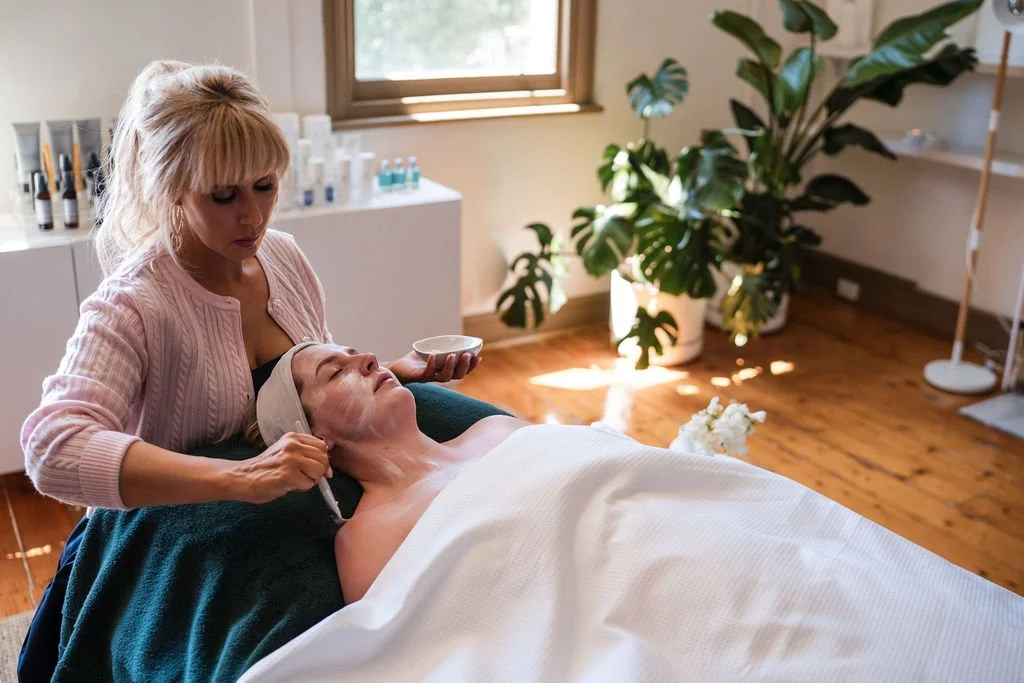 A woman receives a facial treatment at a spa or skincare clinic, lying peacefully with her eyes closed. The spa professional is applying a skincare product or mask on her face in a calm, well-lit room with plants and skincare products on a counter.