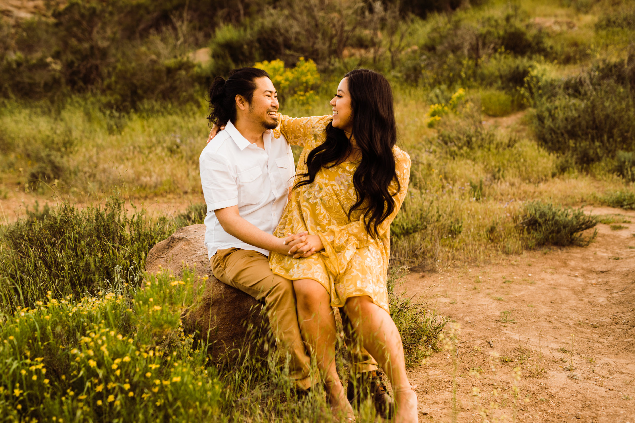 Long-haired man in button-down and khakis, with fiancee in yellow dress on lap | Engagement Outfits | wildflowers at Vasquez Rocks | adventurous elopement photographer Kept Record | www.keptrecord.com