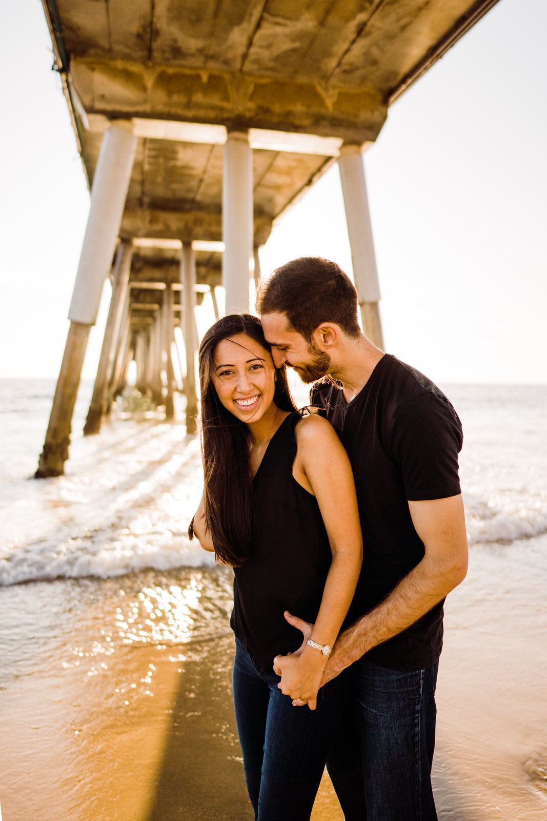 Playful, smiling engaged couple beneath Hermosa Beach Pier