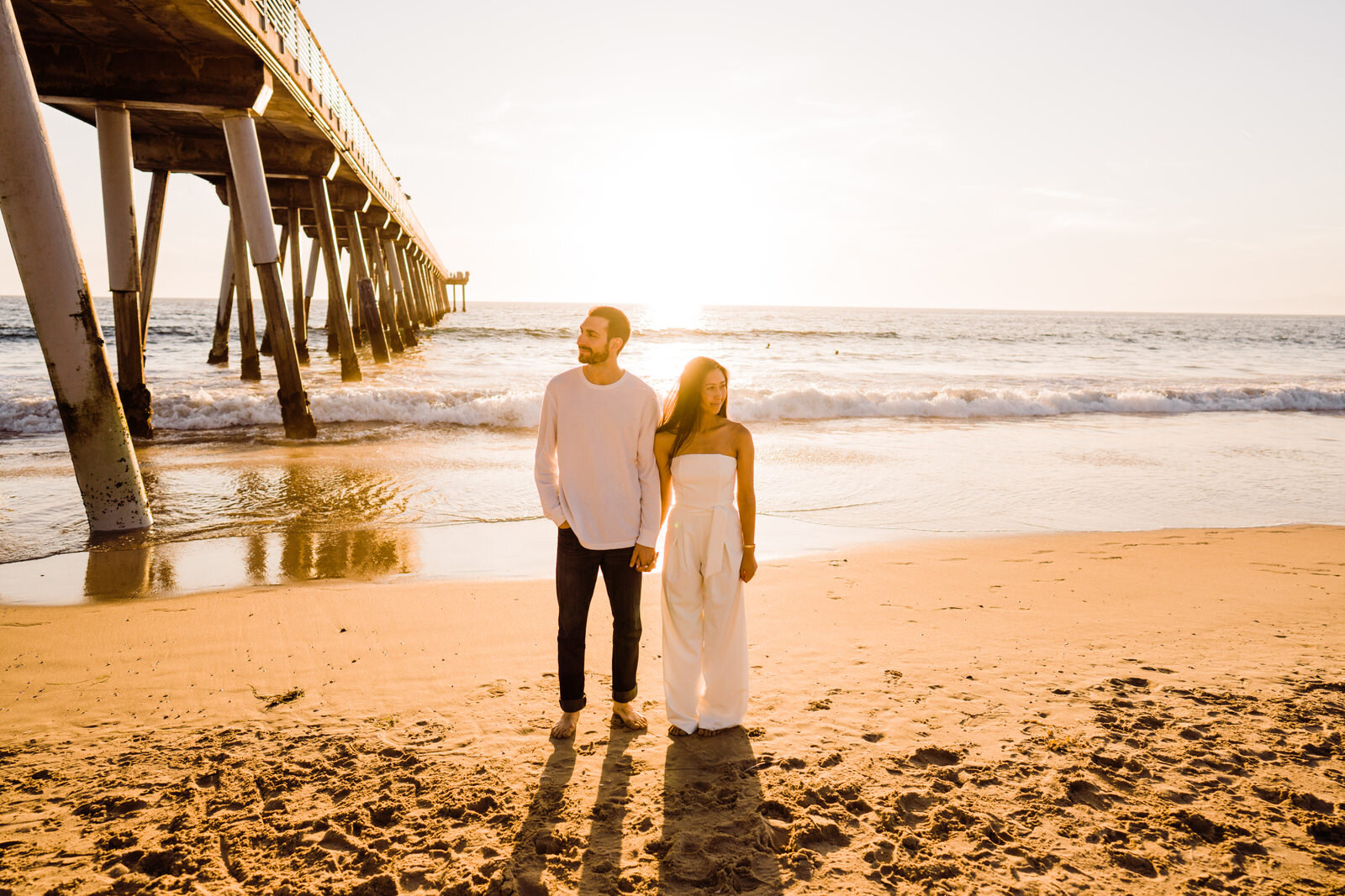 woman wearing white jumpsuit and man wearing white long sleeve and jeans at engagement session at Hermosa Beach