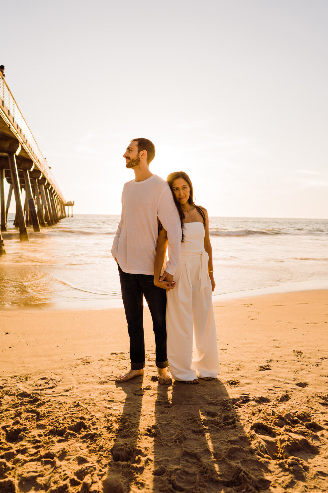 Engaged couple stands back to back at Hermosa Beach Pier wearing white