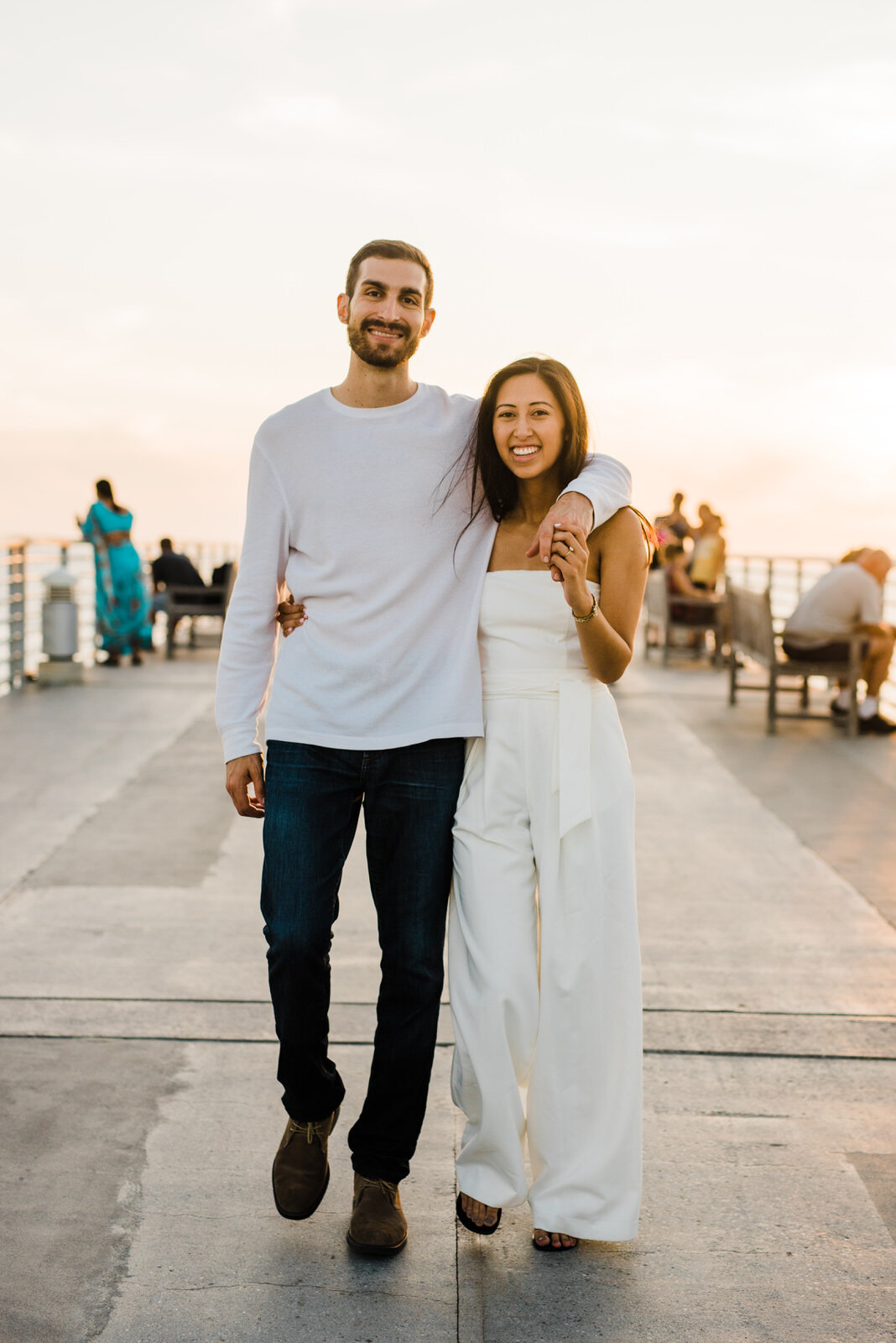 Engaged couple wearing white jumpsuit walks together at Sunset at hermosa beach pier