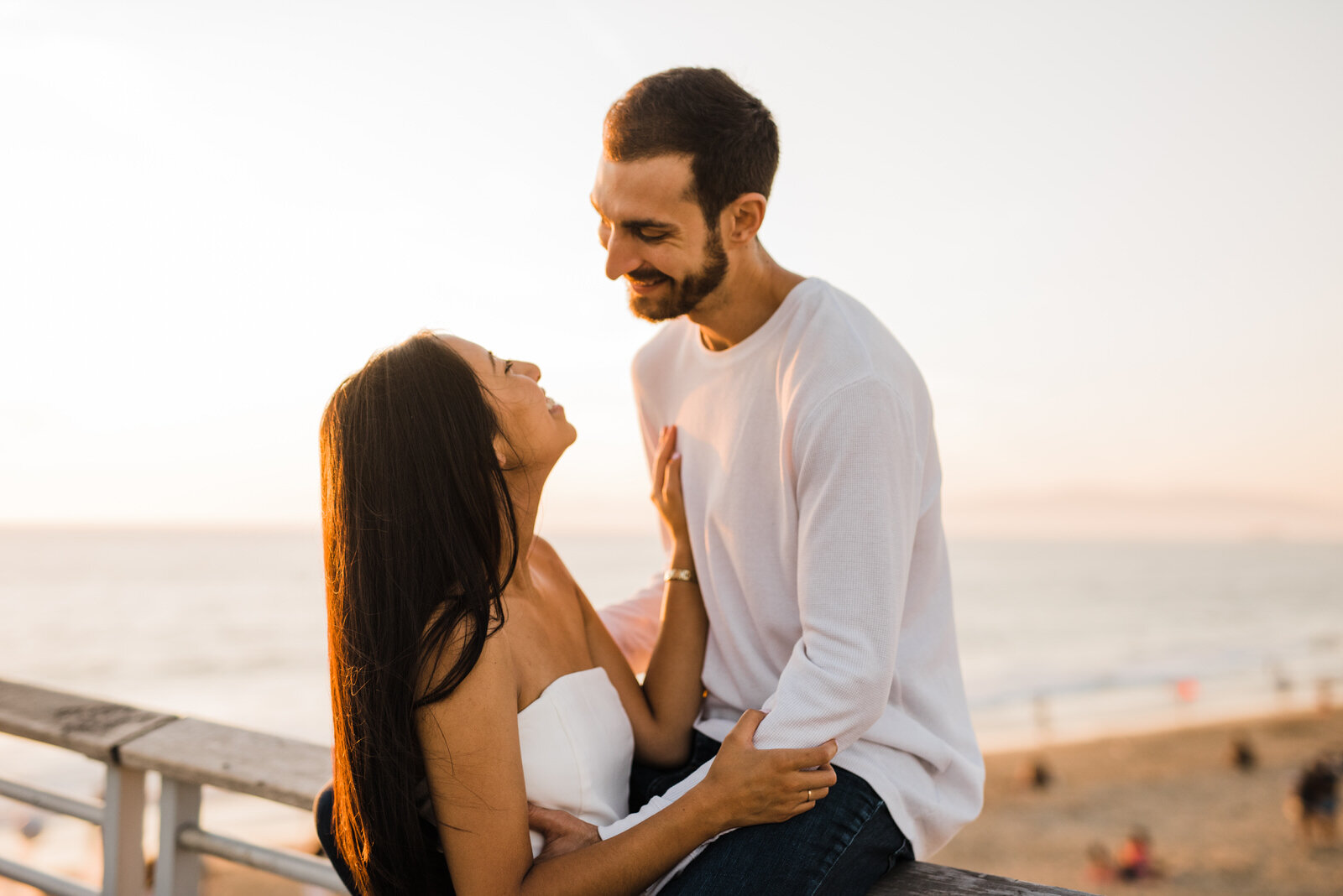 romantic and fun engagement photo with man sitting on railing at Hermosa Beach pier