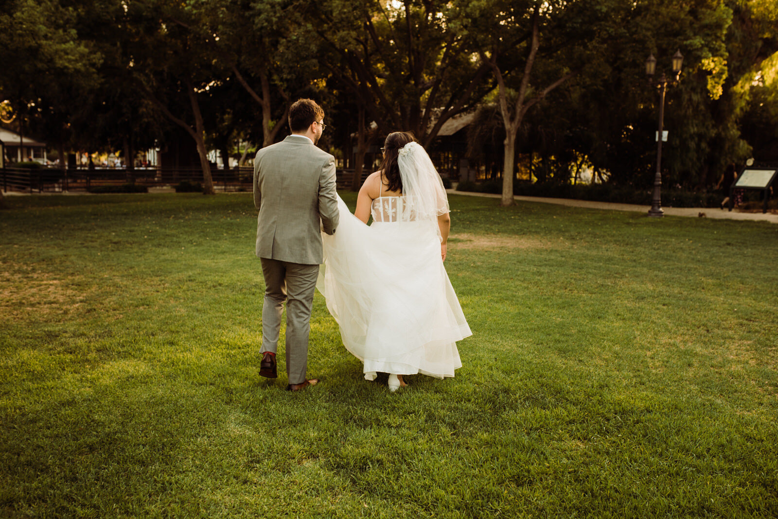 Groom helps carry bride's train as they walk across Heritage Park 