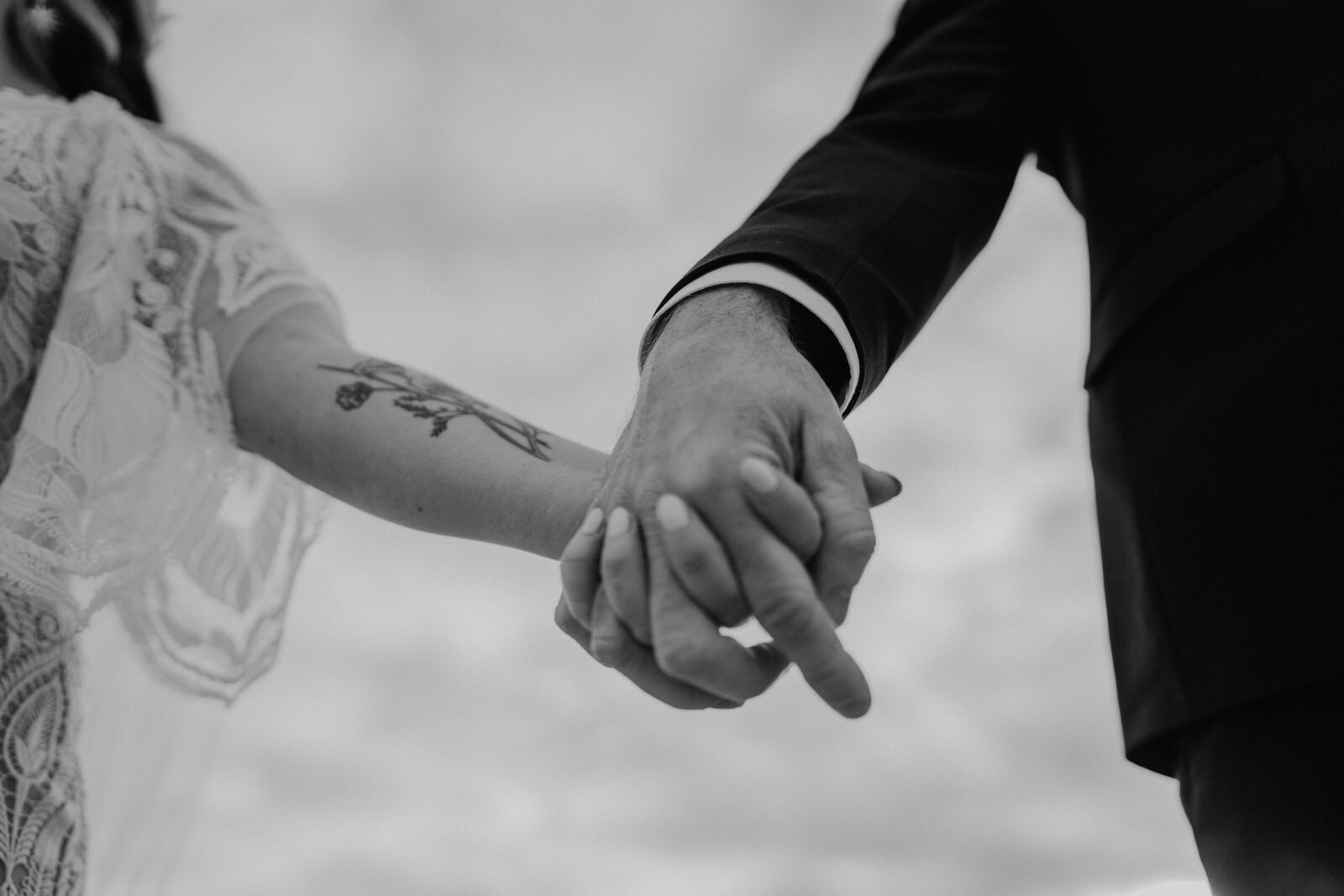 Black and white photo of bride and groom holding hands with bride arm tattoo