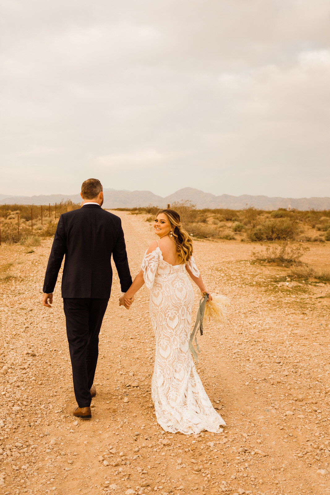 Bride in boho lace gown looks over shoulder walking with groom on dirt road in Las Vegas, NV
