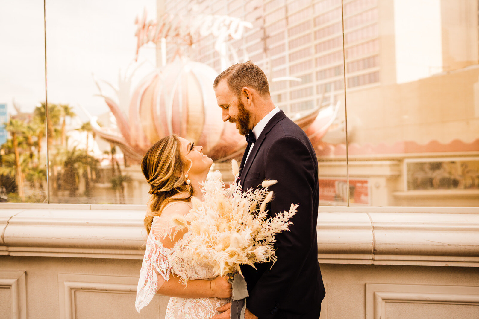Boho bride with pampas grass bouquet and lace dress and groom in navy suit with bowtie in front of The Flamingo Casino on Las Vegas strip