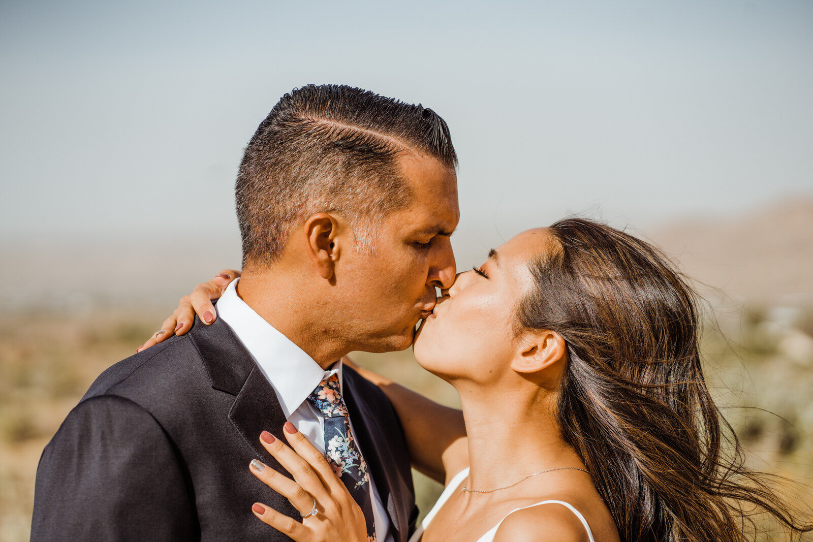 Bride with long black hair kisses grey-haired groom in the sunshine before their wedding ceremony in Joshua Tree