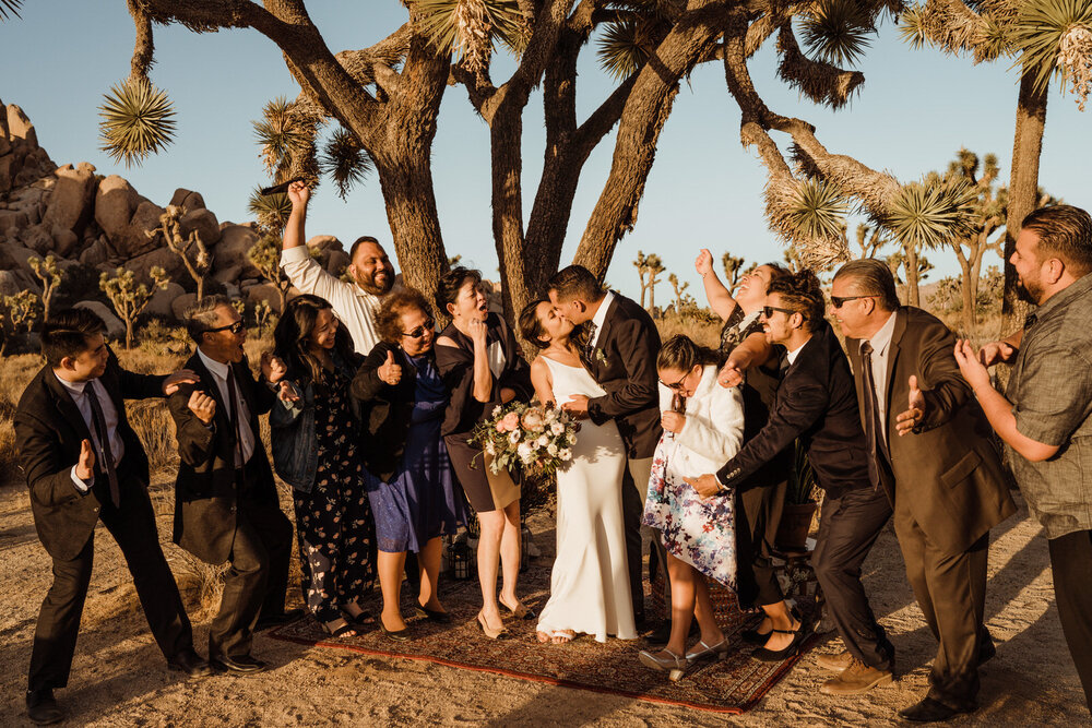 Bride and Groom Kiss beneath a Joshua Tree surrounded by excited, enthusiastic close family members in intimate wedding ceremony 