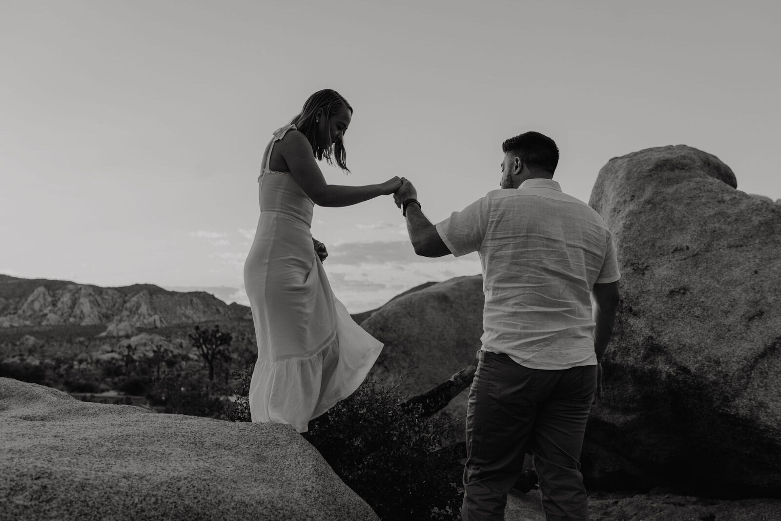 Black and white photo of man helping woman climb off boulder - Sunrise Engagement Session in Joshua Tree, Ca - Photos by Adventurous Elopement + Wedding Photographer Kept Record | www.keptrecord.com