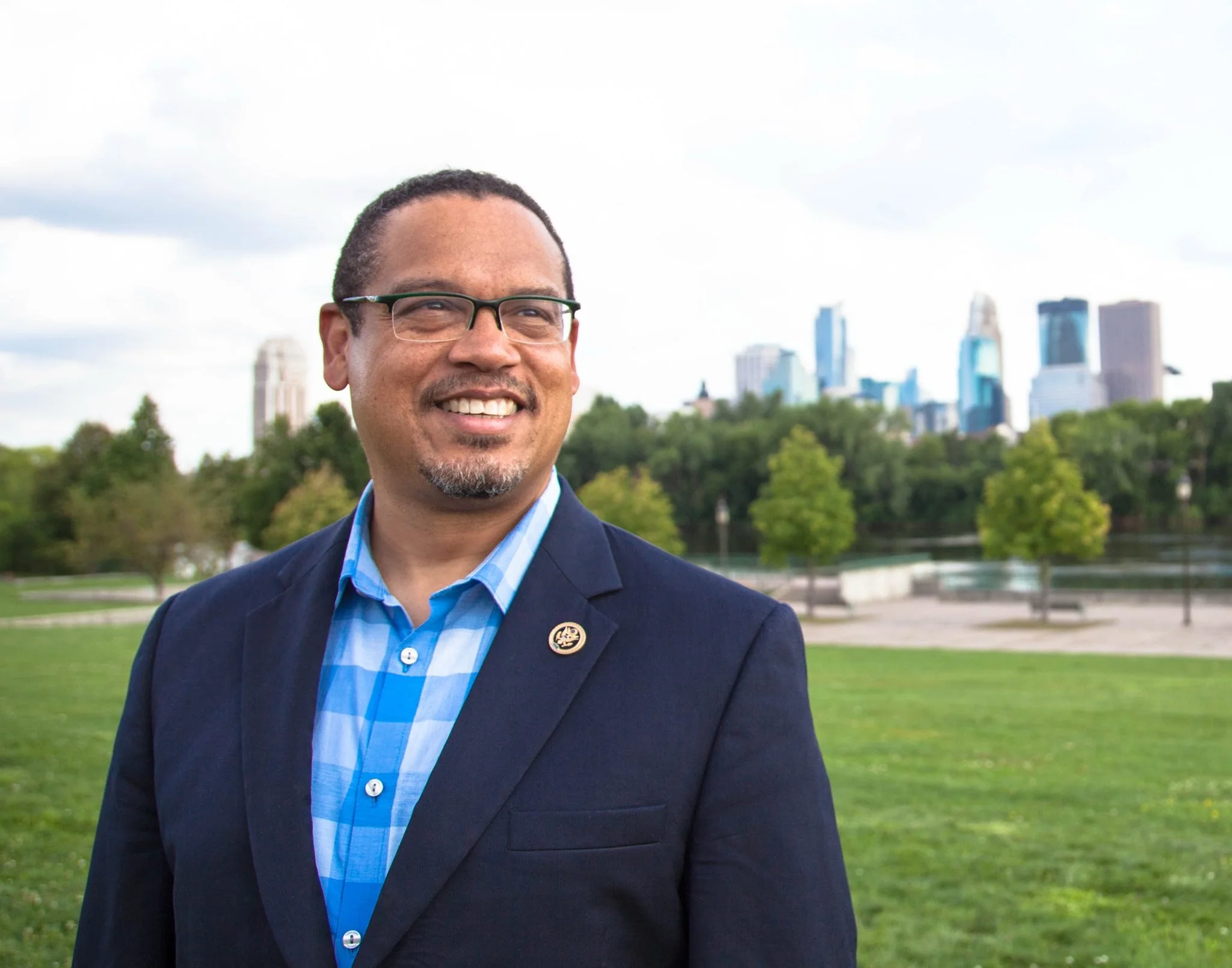 A man smiling outdoors in front of a city skyline with green trees and a park, wearing glasses, a dark blazer, and a blue checkered shirt.