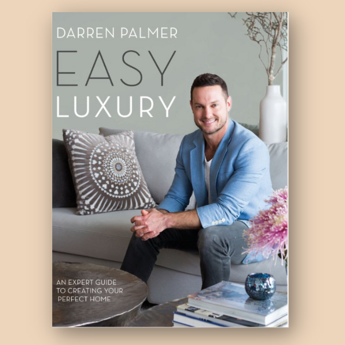 Cover of the book 'Easy Luxury' by Darren Palmer, featuring a man sitting on a sofa with decorative pillows, a vase, and a book on a sleek coffee table. Subtext reads 'An Expert Guide to Creating Your Perfect Home.'
