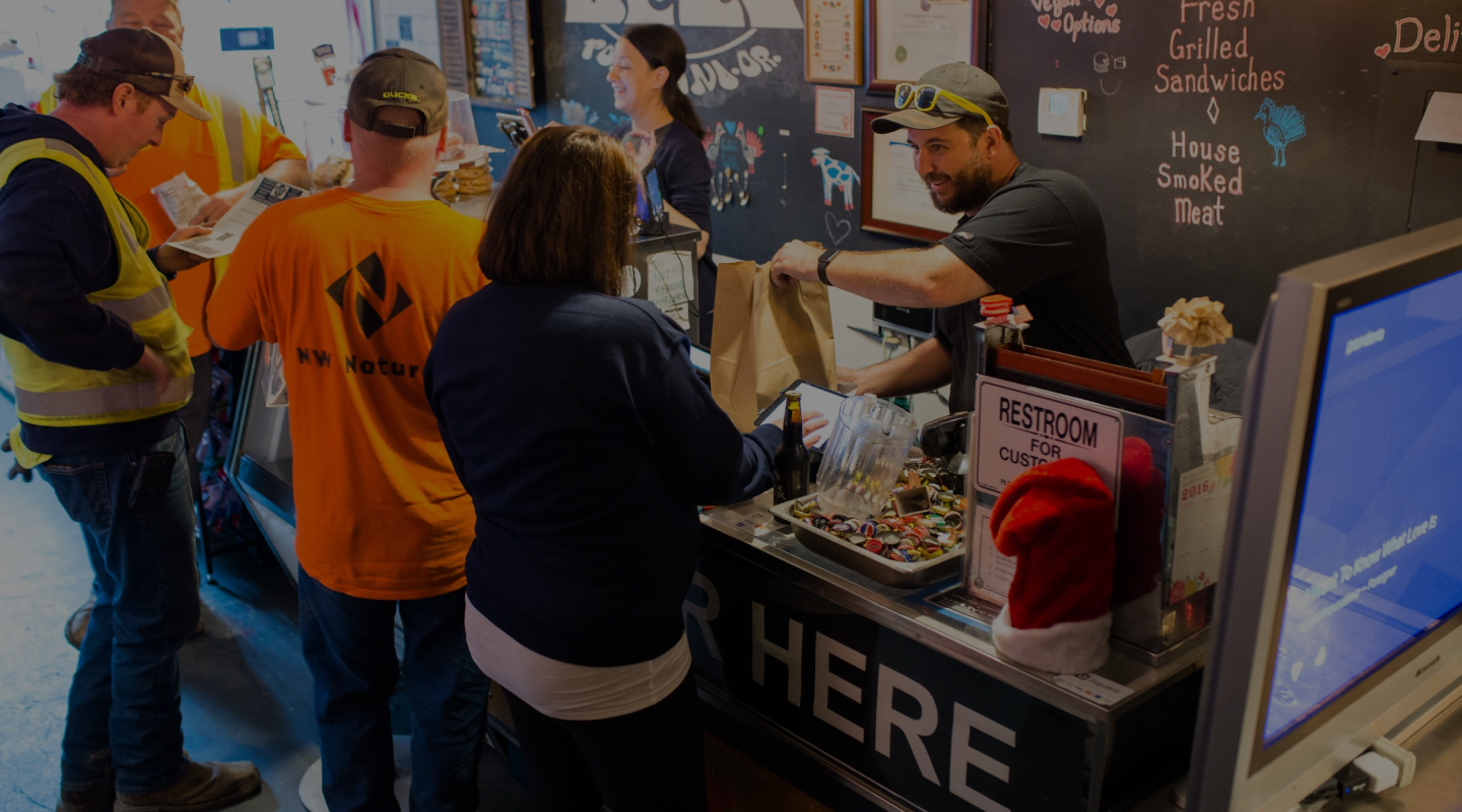 Charlie and Ali handing food across the deli counter, surrounded by neighborhood regulars and the NW Natural crew