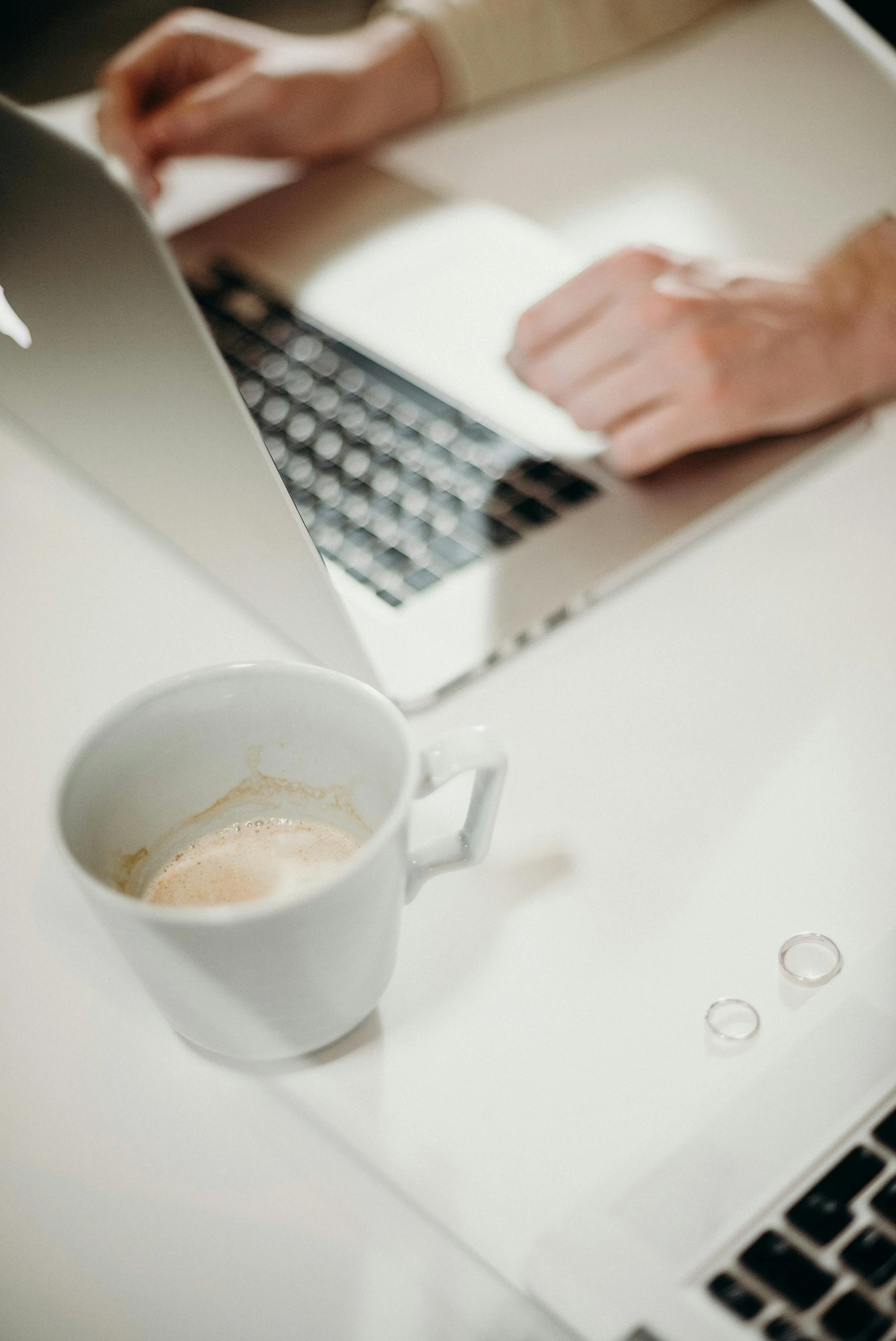 A white coffee mug with a small amount of coffee inside on a white desk, a silver laptop with a person's hands typing, and two small transparent glass rings on the desk.