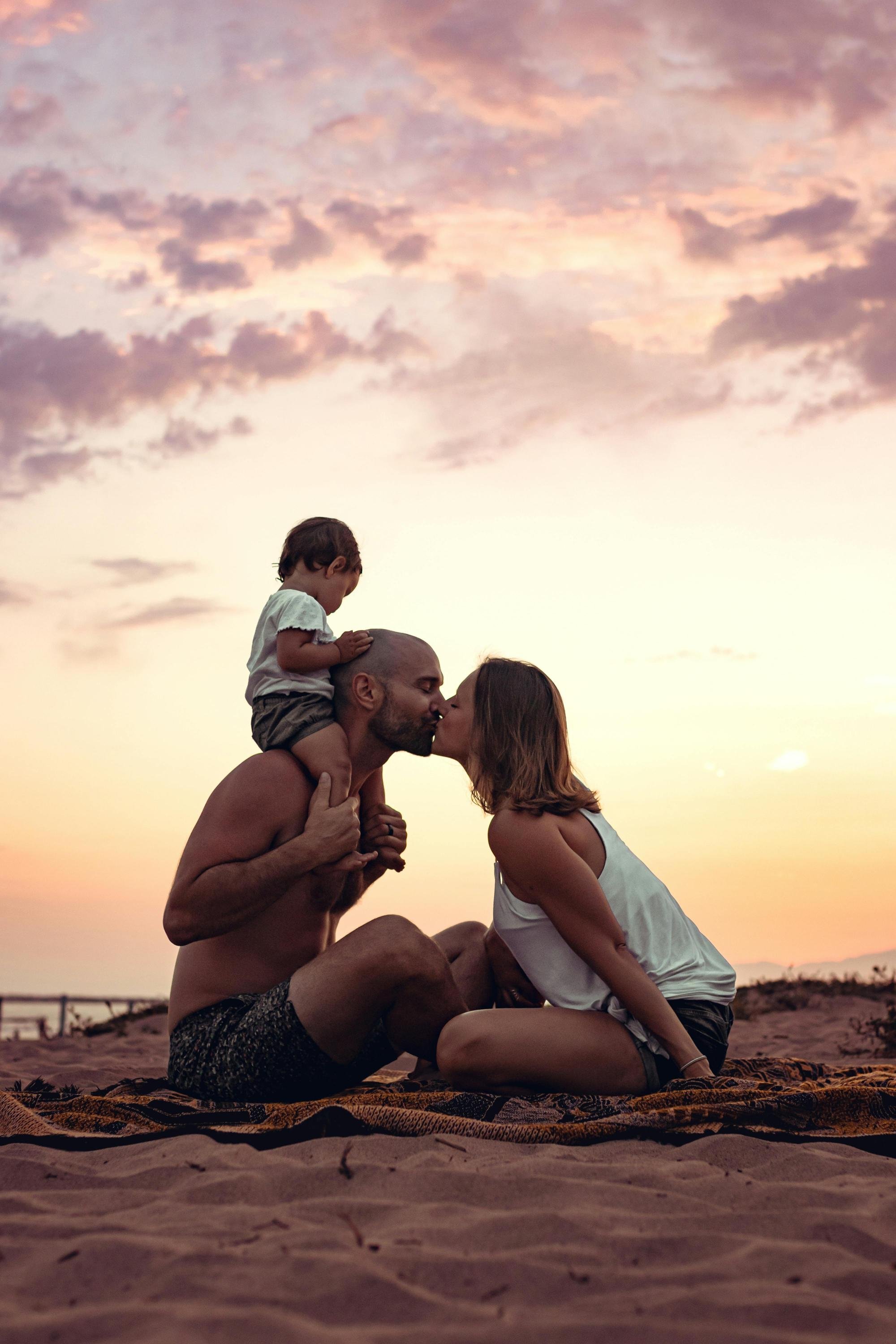 A family of three on a sandy beach during sunset, with the parents kissing and their young child sitting on the father's shoulders.