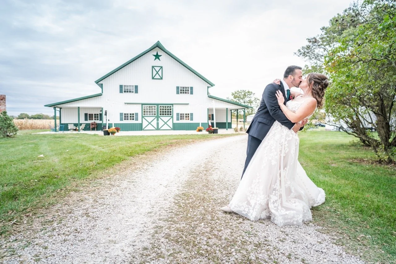 A newlywed couple sharing a kiss on a gravel driveway in front of a barn with white and teal panels, with trees and green grass nearby.