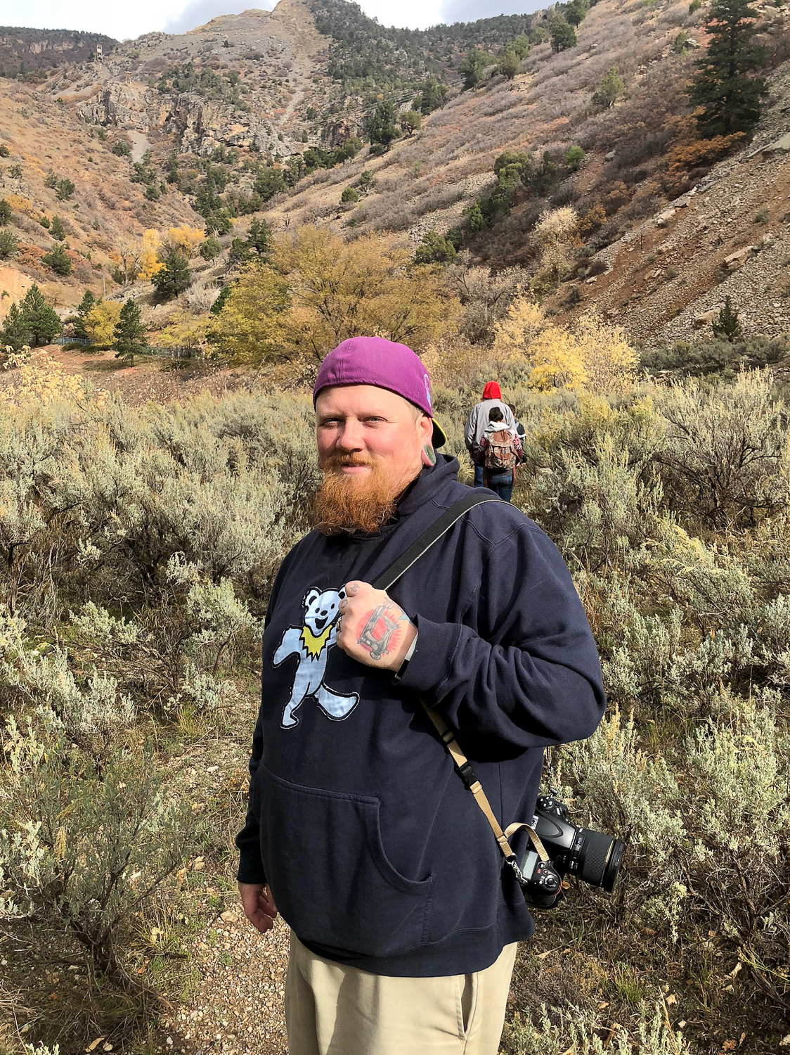 A man with a beard wearing a purple cap backwards, black hoodie with a cartoon bear on it, and beige pants, stands in a desert landscape with mountains and shrubs. He has a camera hanging from his neck and a tattoo on his hand. Two people with backpacks walk behind him on a trail.