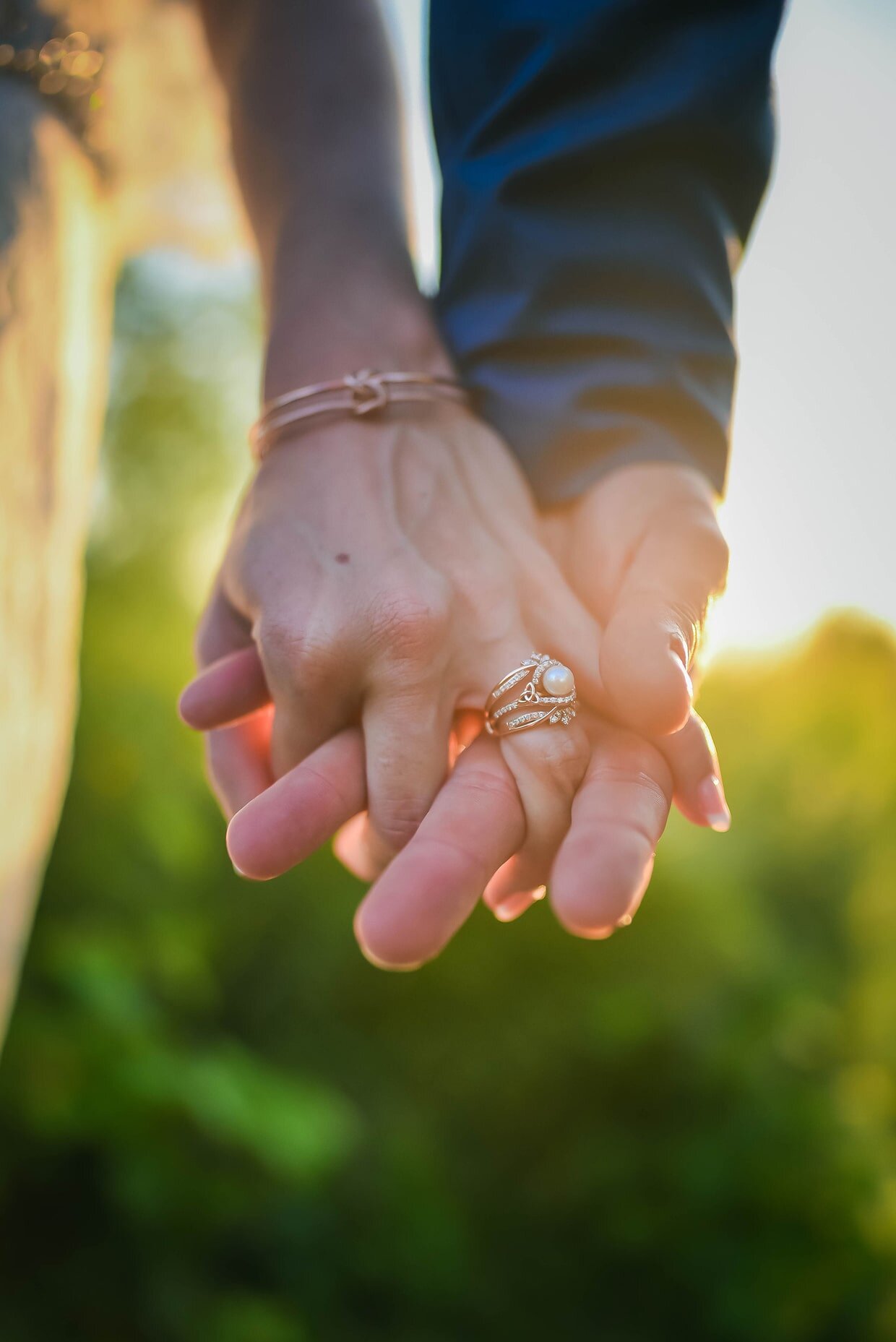 Close-up of two hands holding each other, one with jewelry including rings and an bracelet, with a blurred outdoor background at sunset.