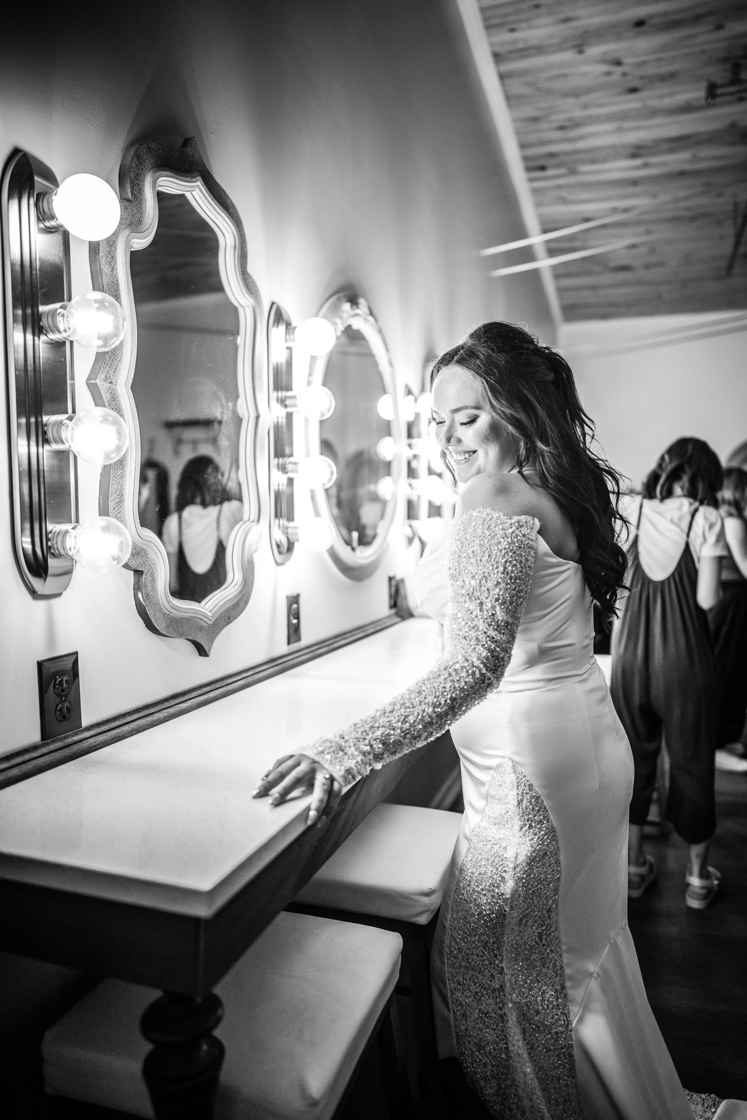 A smiling woman in a wedding dress stands by a counter with illuminated mirrors and light bulbs, in a dressing room.