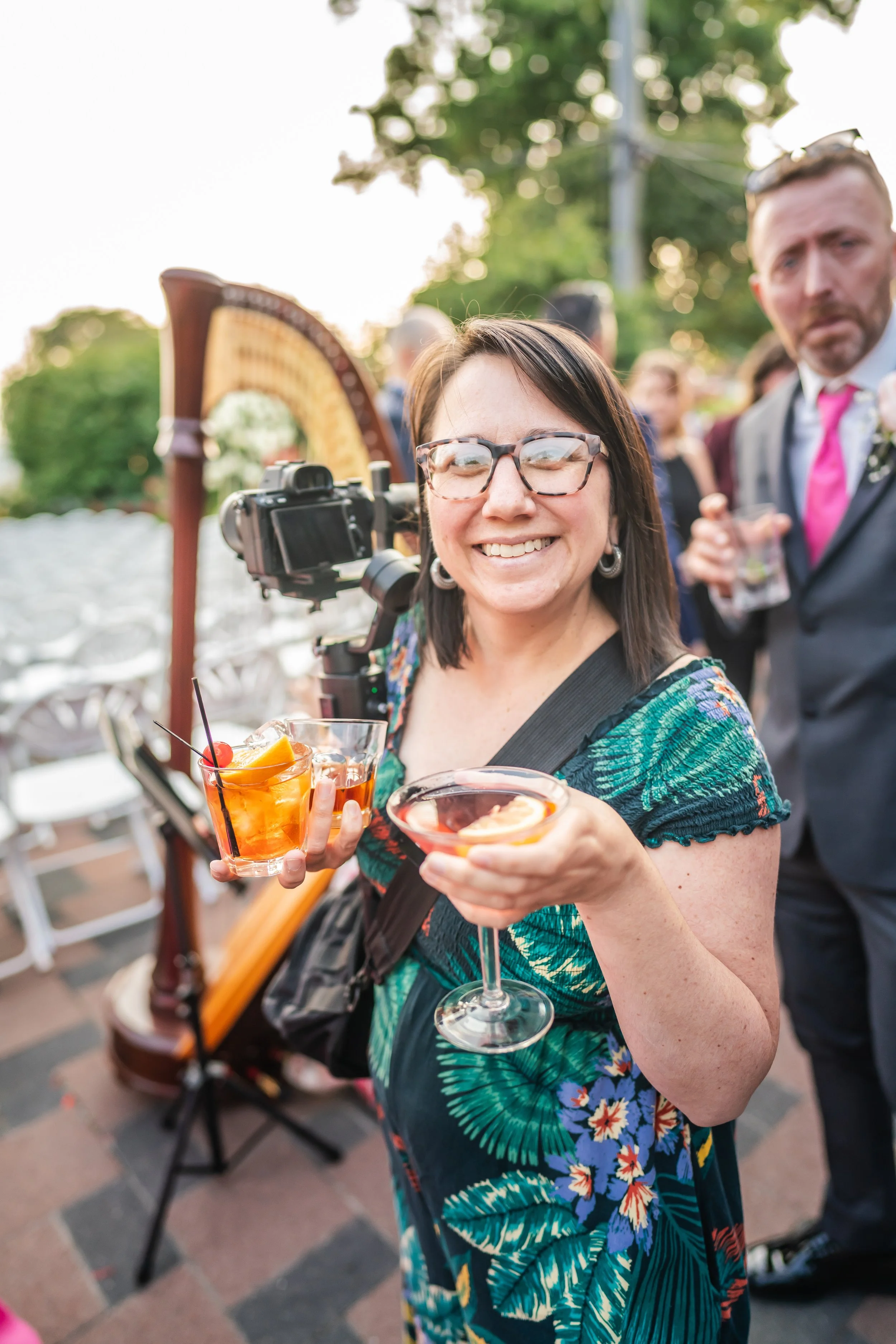 A woman with glasses smiling and holding two cocktails at an outdoor event. She is wearing a colorful dress with a tropical leaf pattern. There are other people and a harp in the background.