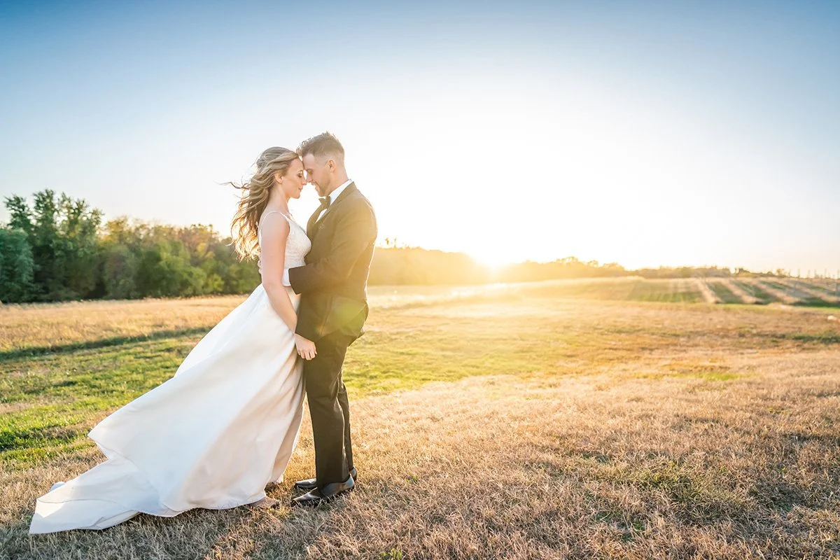 A bride and groom standing close together in an open field at sunset, with trees in the background. The bride is wearing a white wedding gown, and the groom is dressed in a black suit. They are gently touching foreheads, sharing a tender moment.