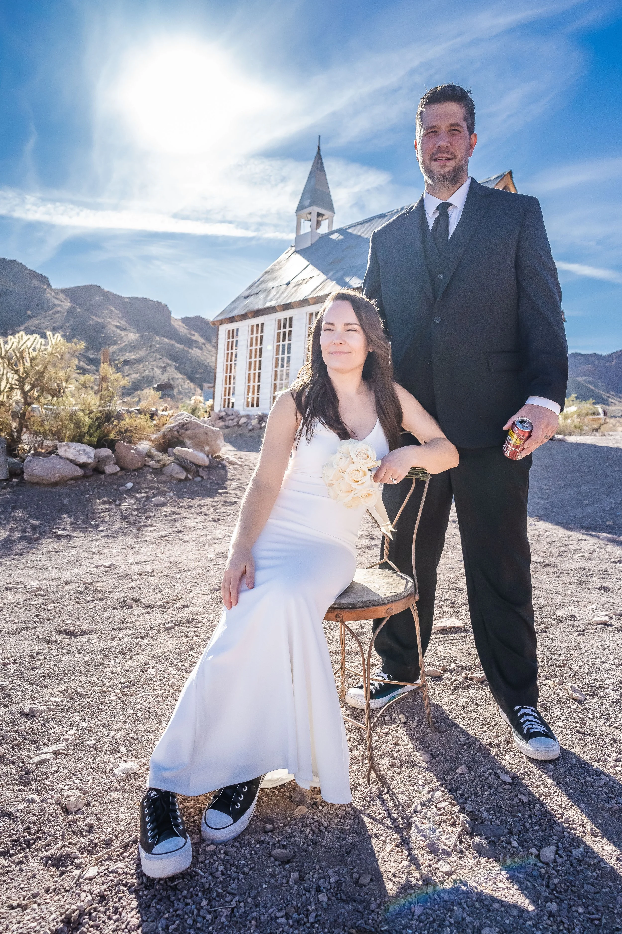 A man in a black suit and tie standing next to a woman in a white dress, who is sitting on a chair with a bouquet of white roses. They are outdoors on rocky terrain with mountains in the background and a small white church behind them. The woman is holding the bouquet and smiling, while the man is looking at the camera with a neutral expression. The bright sun is shining in the clear blue sky.