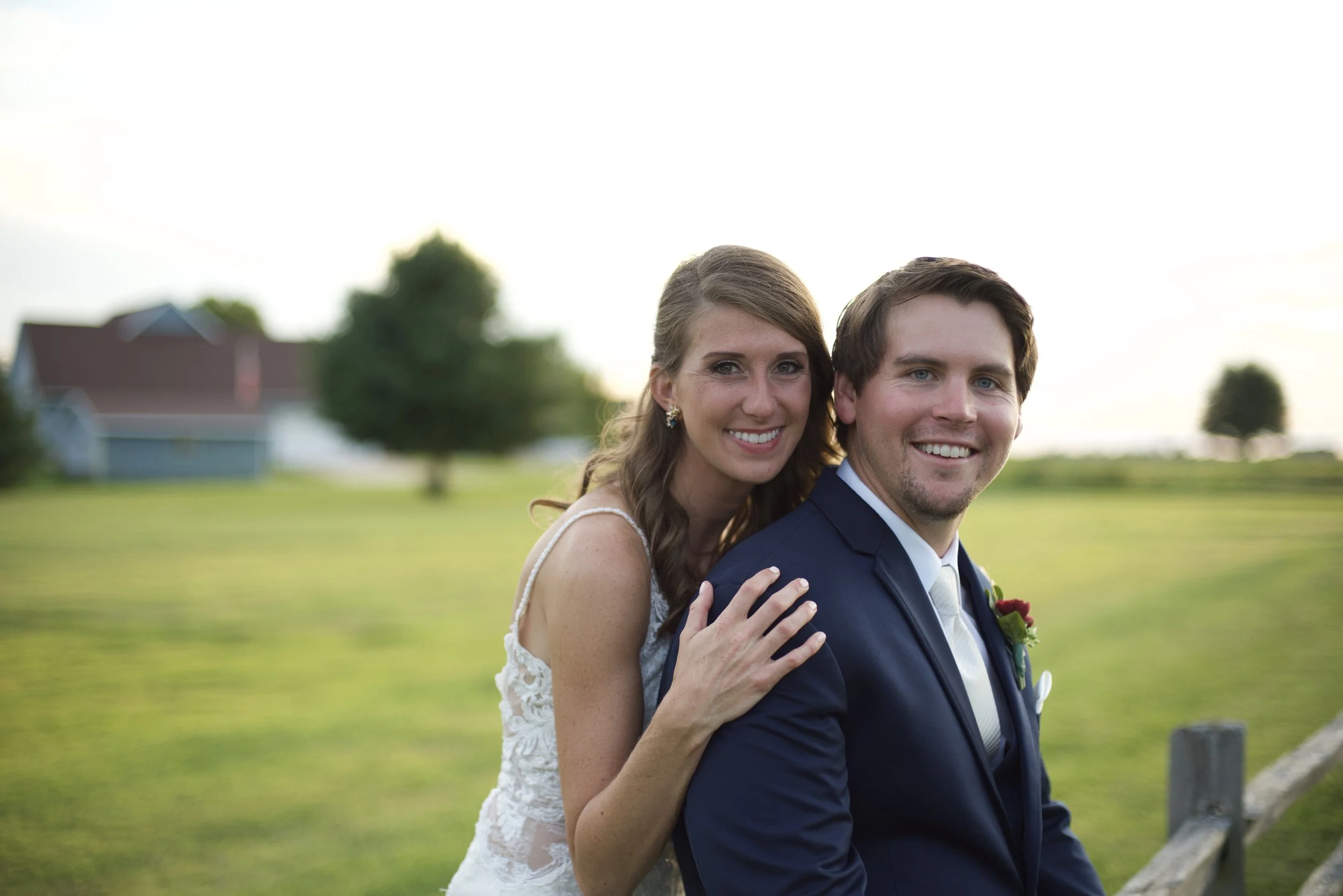 A smiling bride and groom in wedding attire outdoors in a grassy area with trees and a house in the background.