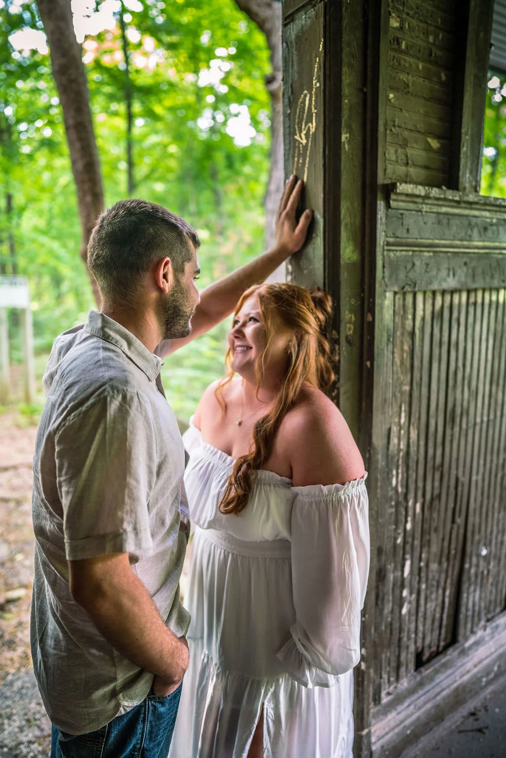A couple sharing a joyful moment outdoors beneath a wooden shelter, with lush green trees in the background. The woman is dressed in a white off-shoulder dress, and the man is wearing a light-colored shirt and jeans. The man has his hand on the woode