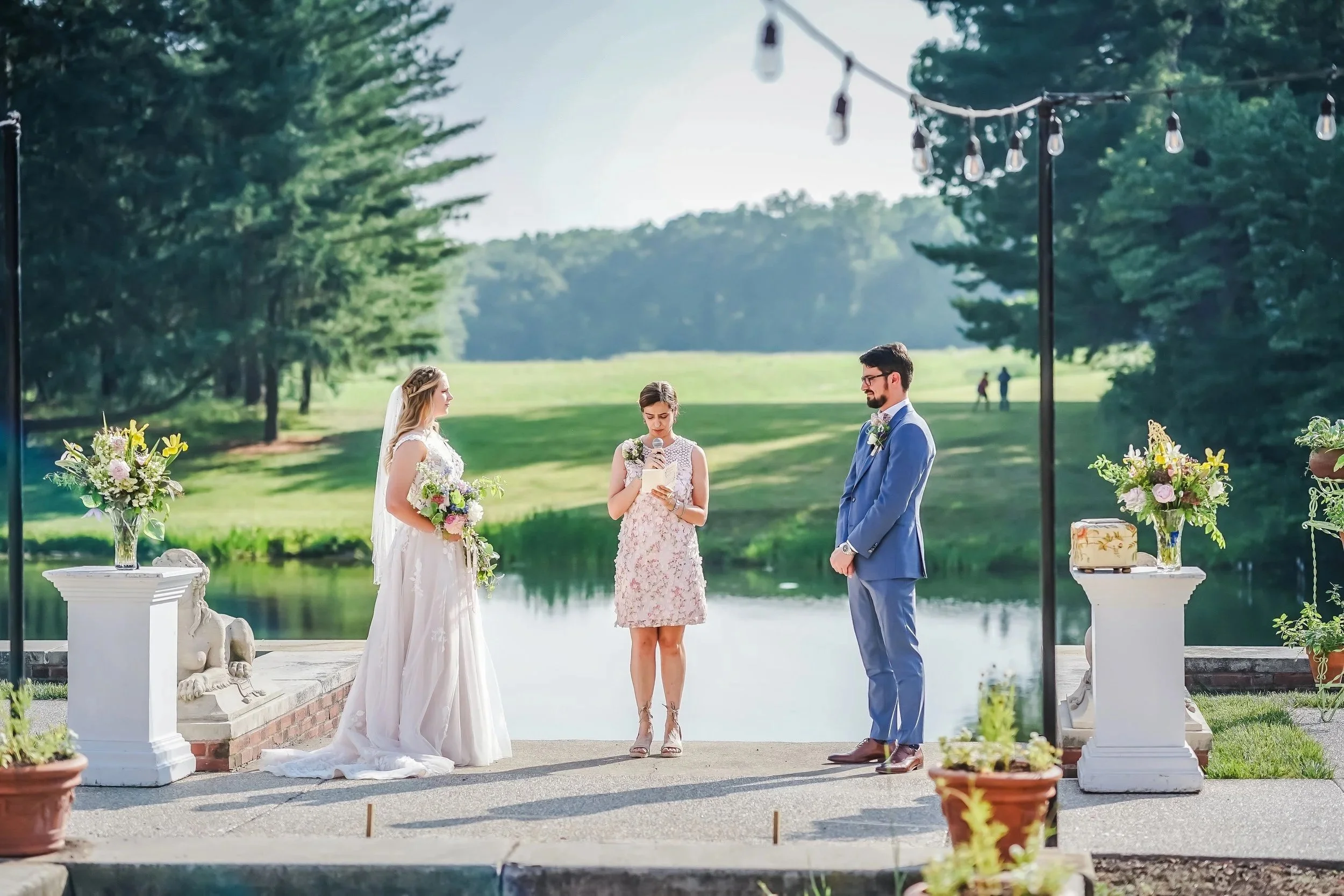 A couple is getting married outdoors by a lake with a person officiating, and the scene is decorated with flowers and string lights.