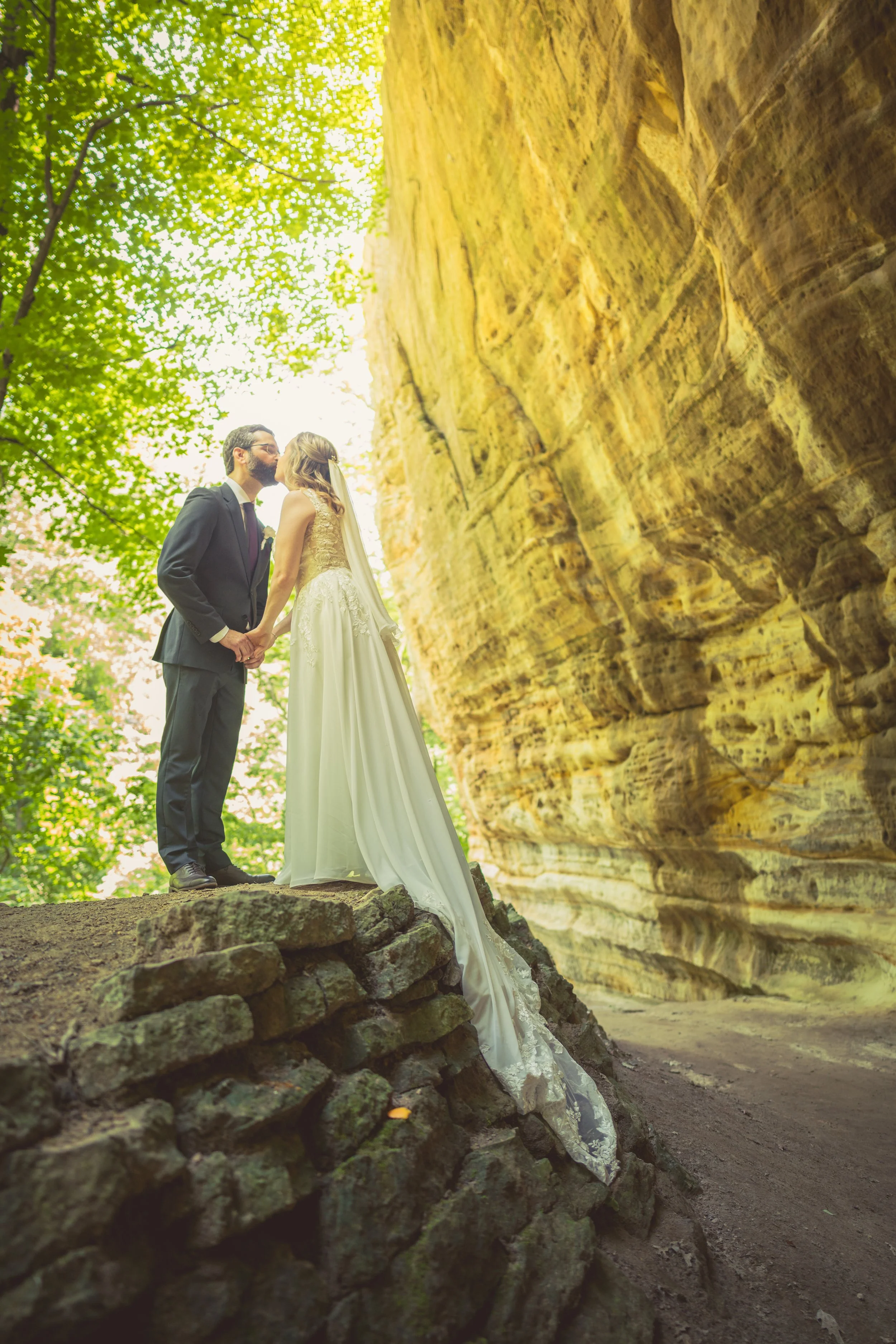 A couple dressed in wedding attire kissing outdoors on a small stone ledge with large rock formations behind them and lush green trees overhead.